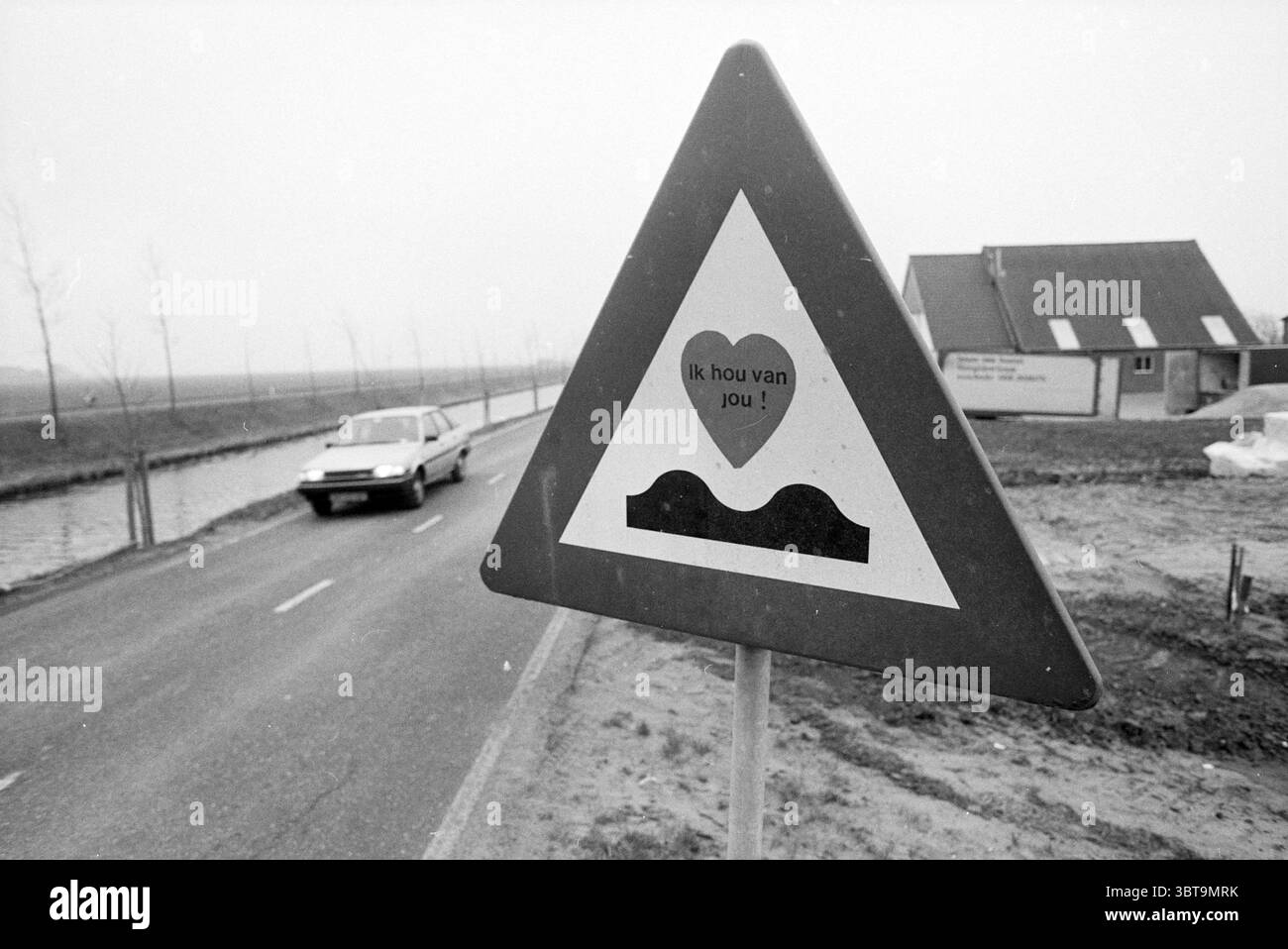Panneau routier indiquant la bosse de vitesse avec autocollant avec le texte I Love You Haarlemmermeer Hoofdweg, Whizgle News, Dutch Desk, The Netherlands, 1950 - 2000 sur 28-01-1992. L'image inclut ces rubriques. La scène présente un contraste saisissant entre les éléments urbains et naturels. Au premier plan, un panneau d'avertissement triangulaire se tient bien en évidence, montrant un symbole de cœur au-dessus d'un graphique qui suggère un creux dans la route. La palette de couleurs est principalement monochromatique, évoquant un sentiment de nostalgie ou une humeur sombre. Sur le côté du panneau, une voiture se déplace sur la route, mettant en évidence une sensation de mouvement. Le véhicule » Banque D'Images