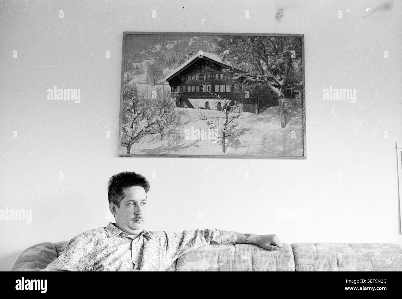 Mr. de Vries Haarlem pays-Bas, Whizgle News, Dutch Desk, pays-Bas, 1950 - 2000 on 03-10-1994. L'image inclut ces rubriques. Dans un espace intérieur légèrement éclairé, un homme est assis confortablement sur un canapé à motifs, son corps légèrement tourné alors qu'il regarde soigneusement sur le côté. Il a les cheveux courts et foncés, et une moustache subtile qui ajoute à son apparence contemplative. La chemise qu'il porte présente un motif floral léger, offrant un contraste doux avec les tons doux du canapé. Au-dessus de lui, une grande œuvre d'art encadrée attire l'attention, représentant un paysage hivernal serein. La scène illustre un RU Banque D'Images