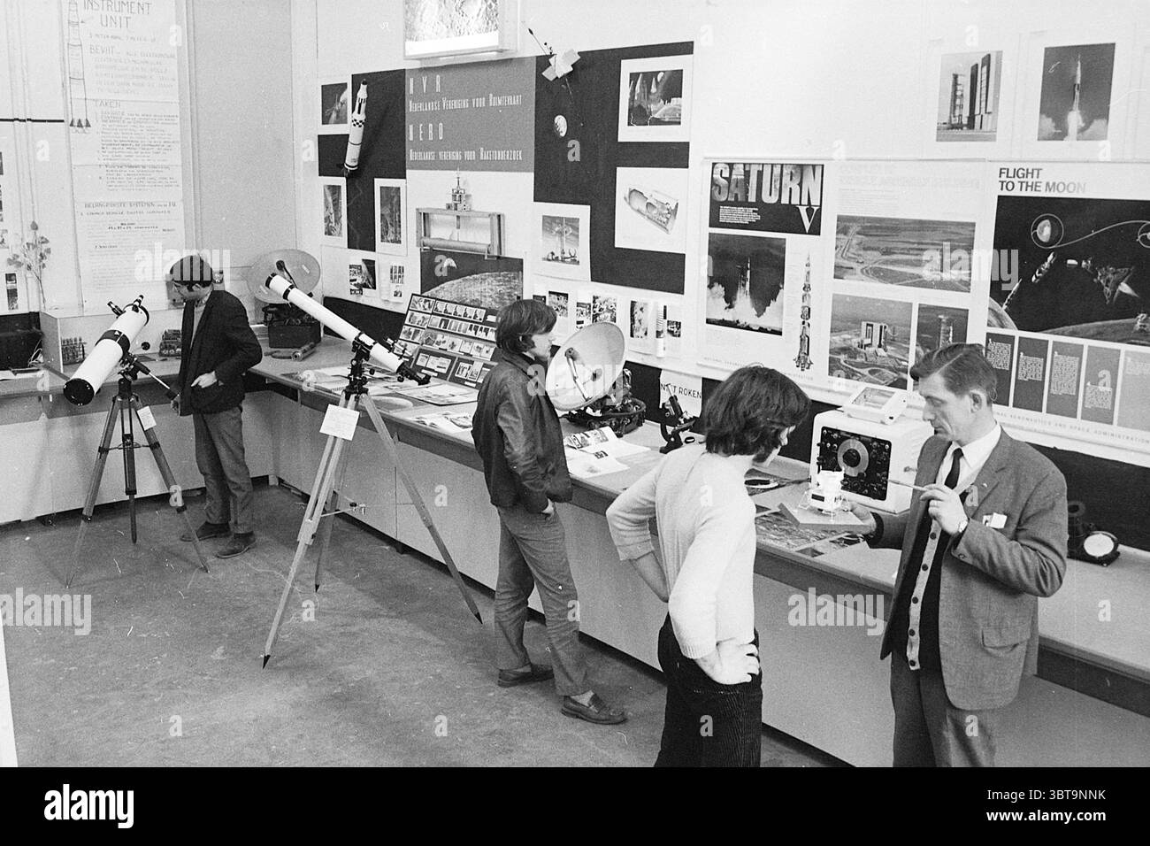 Exposition macro, Whizgle News, Dutch Desk, pays-Bas, 1950 - 2000 sur 26-03-1969. L'image montre ces rubriques. La scène capture un espace d'exposition intérieur animé, caractérisé par un mélange d'exploration scientifique et d'engagement du public. Les murs sont ornés de divers affichages liés à l'astronomie et à l'exploration spatiale. Plusieurs télescopes, dont l'un monté sur un trépied, présentent des finitions métalliques élégantes et un verre réfléchissant, suggérant une focalisation sur l'observation céleste. Autour d'eux, les individus sont engagés dans la conversation et l'étude. Un homme, vêtu d'un Banque D'Images