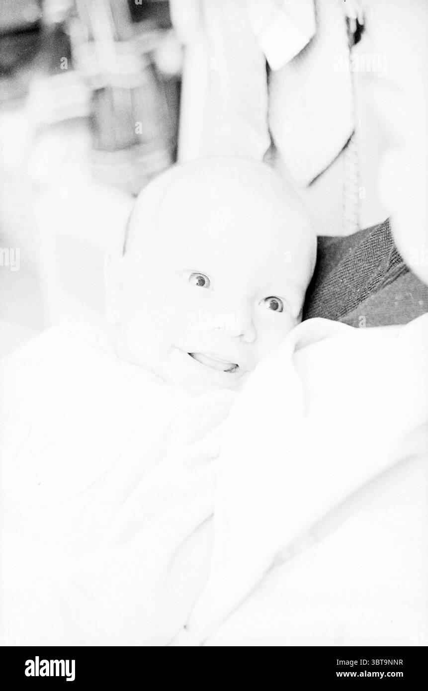 Mère avec bébé dans l'environnement familial., Whizgle News, Dutch Desk, pays-Bas, 1950 - 2000. L'image montre ces rubriques. Dans la scène, un bébé joyeux occupe le premier plan, leurs grands yeux brillants brillants brillent de plaisir. Le bébé, enveloppé confortablement dans une couverture douce et de couleur claire, dégage un sentiment de chaleur et d'innocence. Leur expression est une expression de curiosité et de bonheur, caractérisée par un large sourire qui suggère un moment d’interaction ludique. Autour du bébé est un fond doux et flou qui contient des notes d'autres figures et objets, mais ils restent indistincts, permettant au bébé Banque D'Images