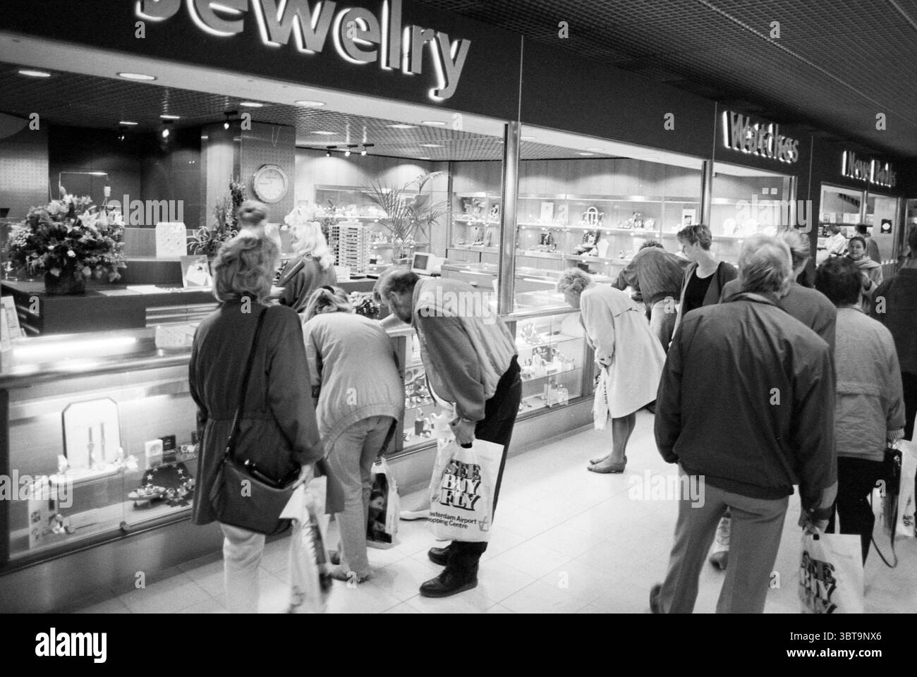 Journée environnementale au terminal de West Schiphol Schiphol, Whizgle News, Dutch Desk, pays-Bas, 1950 - 2000 le 14-05-1993. L'image inclut ces rubriques. La scène capture une bijouterie animée, où une foule de clients observe attentivement les étalages. Divers individus sont rassemblés devant une vaste vitrine en verre remplie de bijoux éblouissants, montrant un mélange de bagues, colliers et boucles d'oreilles étincelants. Les gens dans la scène sont divers en âge et en tenue ; certains sont habillés de façon décontractée tandis que d'autres présentent un style plus poli, suggérant qu'ils sont soit des fenêtres s Banque D'Images