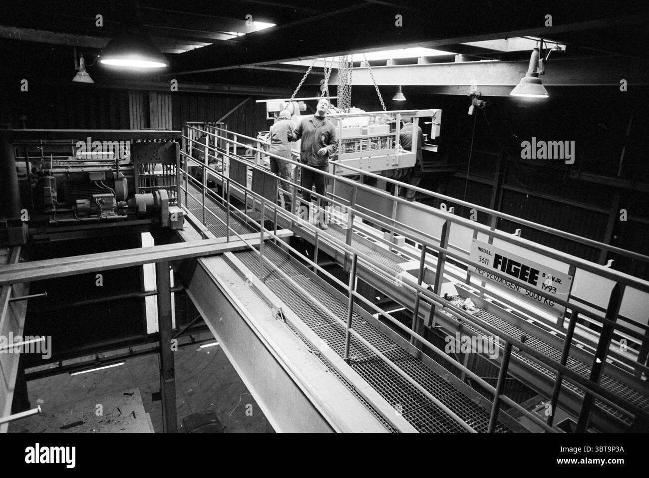 Installation d'une grue à VAM H'lem Haarlem pays-Bas, Whizgle News, Dutch Desk, pays-Bas, 1950 - 2000 le 14-01-1993. L'image contient ces rubriques. La scène capture un environnement industriel caractérisé par ses caractéristiques robustes. Au-dessus, un plafond sombre orné de quelques plafonniers projette une lueur vive, éclairant la scène dans des tons discrets. L'éclairage crée un contraste entre les zones bien éclairées et les ombres plus profondes qui se cachent dans les coins, contribuant à une ambiance un peu gritty. Au premier plan, deux travailleurs sont engagés dans la manipulation d'un grand objet suspendu, suggérant Banque D'Images