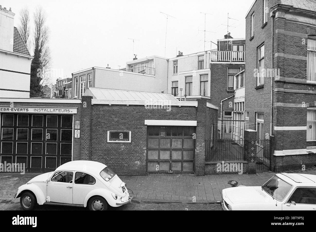 Bâtiments et fissures Schotersingel 117 Huizen Haarlem Haarlem Schotersingel pays-Bas, Whizgle News, Dutch Desk, pays-Bas, 1950 - 2000 on 27-01-1977. L'image contient ces rubriques. La scène capture un cadre urbain calme caractérisé par un mélange d'architecture résidentielle et commerciale. À gauche, un bâtiment en brique présente de grandes fenêtres en verre affichant une série de carrés sombres, indiquant peut-être une entrée ou un magasin. Sa façade est ornée d'une série de colonnes cannelées et d'un toit légèrement en surplomb, lui donnant un look classique dans un décor plus contemporain. Au premier plan Banque D'Images