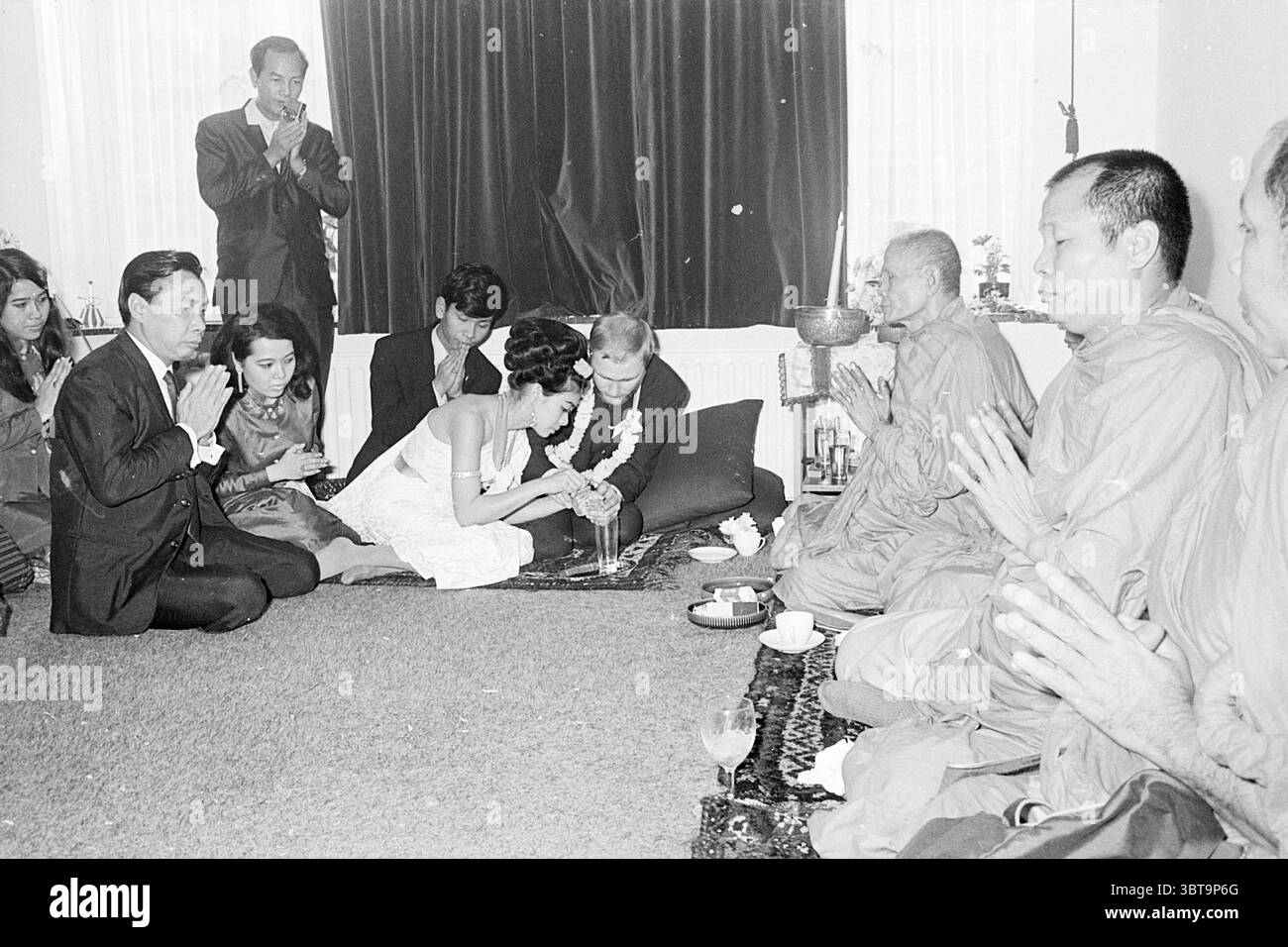 Mariage bouddhiste avec grand prêtre de Thaïlande Heemstede pays-Bas, Whizgle News, Dutch Desk, pays-Bas, 1950 - 2000 on 10-11-1968. Ces rubriques sont illustrées dans l'image. La scène se déroule dans une salle spacieuse et chaleureusement éclairée, où se déroule une cérémonie solennelle mais joyeuse. Au premier plan, un couple s'agenouille étroitement ensemble, se concentrant intensément sur un rituel. La femme, vêtue d'une élégante robe blanche, est ornée de guirlandes florales, ses cheveux coiffés dans un updo complexe. L'homme à côté d'elle est dans un costume sombre, les deux montrant des expressions de vénération et d'intimité. Autour d'eux sont indiv Banque D'Images