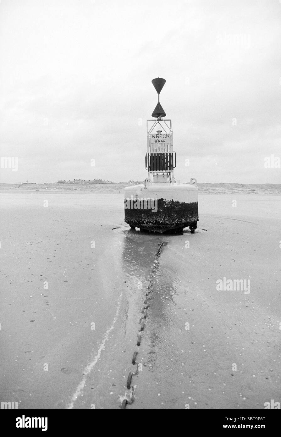 Bouée échouée IJmuiden IJmuiden pays-Bas, Whizgle News, Dutch Desk, pays-Bas, 1950 - 2000 le 25-01-1993. L'image montre ces rubriques. La scène présente un paysage monochromatique marqué dominé par une bouée solitaire positionnée au centre. La bouée, surélevée au-dessus de l'eau, est construite à partir d'un matériau altéré, montrant des signes d'âge et d'usure à travers des taches de rouille et de peinture décolorée. Au sommet de la bouée, il y a un marquage distinctif avec un symbole de flèche, indiquant le guidage de navigation. Entourant la bouée, l'eau semble calme mais reflète le ciel couvert, créant un s. Banque D'Images