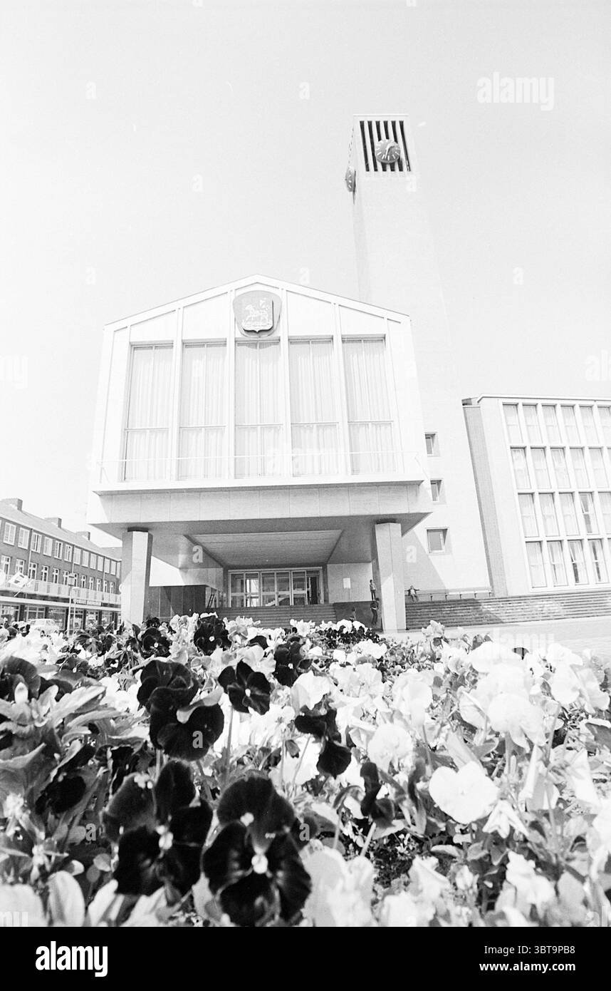 Mairie avec tour IJmuiden (municipalité de Velsen) extérieur IJmuiden plein 1945 pays-Bas, Whizgle News, Dutch Desk, pays-Bas, 1950 - 2000 on 15-05-1974. L'image inclut ces rubriques. La scène présente une structure architecturale moderne caractérisée par des lignes épurées et un design minimaliste, affichant en évidence un clocher. La façade du bâtiment est majoritairement blanche, ce qui lui confère un aspect élégant et contemporain. À la base de la structure, une variété de fleurs vibrantes, y compris des nuances de mauves et de pansies blanches, créent un contraste saisissant avec la lumière du bâtiment Banque D'Images