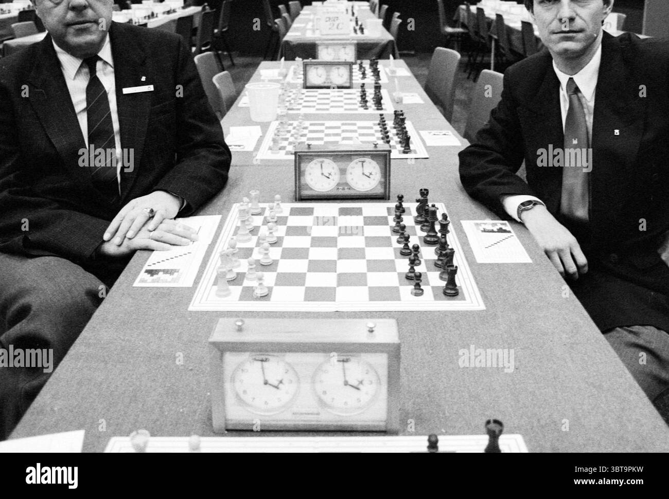 Ancien et nouveau trésorier TVH, Whizgle News, Dutch Desk, pays-Bas, 1950 - 2000 au 25-01-1990. Voici les rubriques de l'image. La scène capture un moment d'un tournoi d'échecs se déroulant dans une salle spacieuse et organisée remplie de nombreuses tables. À l'avant-garde, deux hommes sont assis à une table, chacun concentré sur leur échiquier doublé de pièces positionnées pour un match en cours. La table affiche un échiquier à carreaux avec des carrés sombres et clairs alternant, et les pièces sont placées dans leurs positions de départ, principalement en noir et blanc. À proximité, deux horloges d'échecs classiques sont bien en évidence, leur visage Banque D'Images
