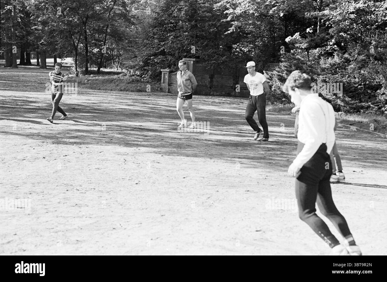 DIRECTEURS INFORMATIQUES de formation au patinage à sec. Patinage, Whizgle News, Dutch Desk, pays-Bas, 1950 - 2000 le 09-10-1965. L'image contient ces rubriques. Dans un cadre extérieur ensoleillé, un groupe de quatre personnes est engagé dans un jeu décontracté, créant une atmosphère animée. Le sol, sablonneux et légèrement inégal, suggère un espace récréatif bien utilisé, peut-être un champ ou une zone de parc. La scène est remplie de diverses nuances de vert provenant des arbres environnants, offrant une toile de fond luxuriante. La lumière du soleil filtre à travers les feuilles, projetant des ombres étouffées sur le sol. Parmi les participants, deux sont des hommes et deux sont des femmes, Banque D'Images