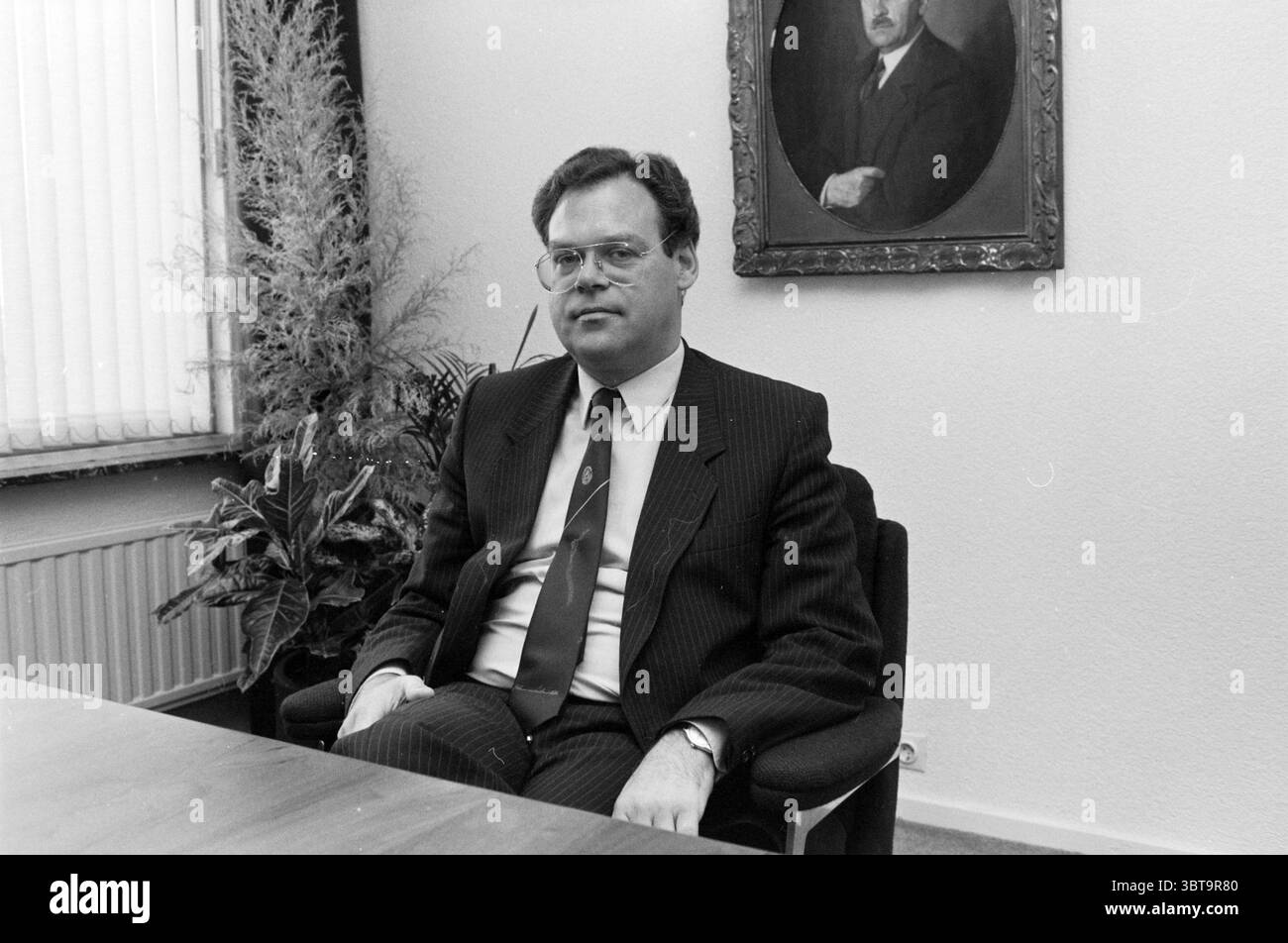 Dir. Droste Mr de Vries Droste, Whizgle News, Dutch Desk, pays-Bas, 1950 - 2000 on 21-04-1985. Ces rubriques sont illustrées dans l'image. Dans un cadre stérile et professionnel, un homme est assis à une simple table en bois, dégageant un air de formalité. Il est habillé d'un costume sombre à fines rayures, accompagné d'une chemise blanche et d'une cravate, qui ajoute une touche de couleur, probablement en bleu foncé ou rouge. Ses lunettes reposent bas sur son nez, et il regarde directement devant lui avec une expression sérieuse, suggérant un comportement contemplatif ou professionnel. Derrière lui, le mur est orné d'un grand portrait encadré d'un homme, Banque D'Images