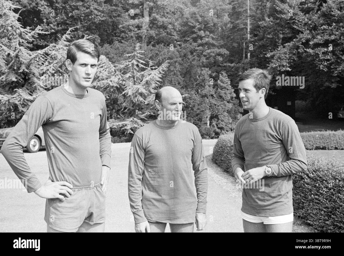 3 hommes en uniforme sportif, Whizgle News, Dutch Desk, pays-Bas, 1950 - 2000 le 02-08-1969. L'image inclut ces rubriques. La scène met en scène trois individus debout ensemble à l'extérieur, sur fond de verdure luxuriante et dense. Les grands arbres, avec leur riche feuillage, créent un cadre naturel autour du trio, contribuant à une atmosphère sereine. Les gens sont habillés dans des hauts de sport à manches longues assortis, mettant l'accent sur un thème cohérent. Les vêtements sont probablement dans une nuance sourde, peut-être gris ou bleu clair, complétant l'environnement tranquille. Le premier individu, notamment plus grand, stan Banque D'Images