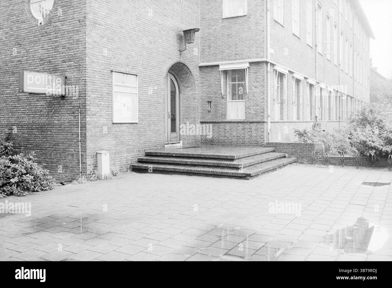 Entrée au poste de police IJmuiden police IJmuiden pays-Bas, Whizgle News, Dutch Desk, pays-Bas, 1950 - 2000 on 13-08-1987. Ce sont les éléments de l'image. La scène représente l'entrée d'un bâtiment caractérisé par sa façade en briques, qui présente une couleur brun rougeâtre sourde. L'architecture a un design utilitaire, mettant en vedette plusieurs grandes fenêtres qui permettent à la lumière naturelle d'éclairer l'intérieur. Au premier plan, il y a une série de marches menant à une arche arrondie qui sert d'entrée principale. Les marches sont faites d'une pierre plus foncée, complétée par le Banque D'Images