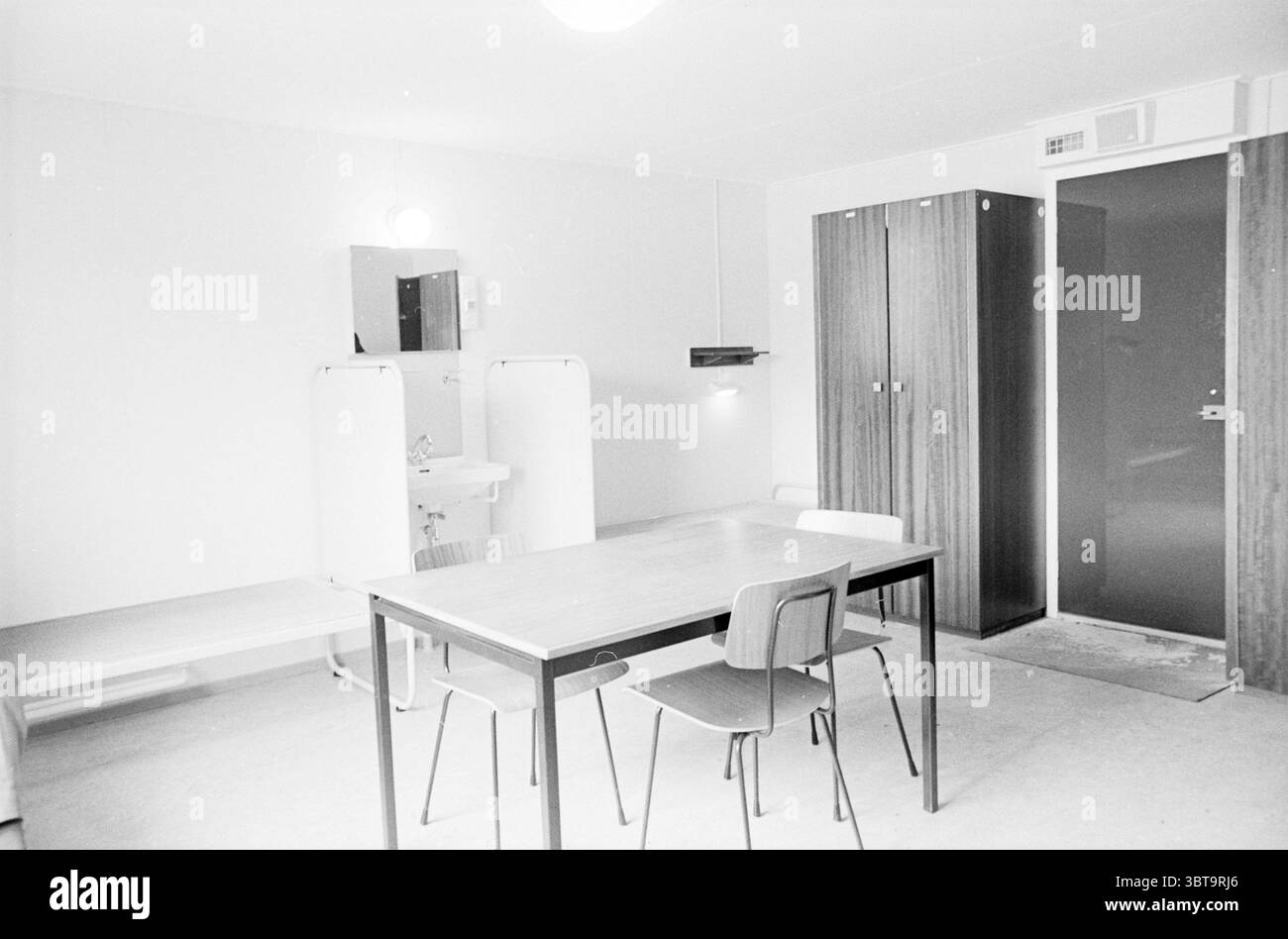 Ext. Et intérieur appartement de nouvelle construction, Whizgle News, Dutch Desk, pays-Bas, 1950 - 2000 sur 07-1965. Ce sont les éléments de l'image. La scène représente un espace intérieur monochrome qui dégage une esthétique minimaliste. Les murs sont peints d'un blanc doux et neutre, contribuant à une atmosphère propre et ouverte. Un plafonnier circulaire unique est suspendu au-dessus, diffusant une lumière uniforme dans toute la pièce. Au centre, une table rectangulaire simple est entourée de quatre chaises en bois légères. Les chaises ont un design classique avec des jambes minces et des dossiers légèrement incurvés, soulignant un sens de functio Banque D'Images