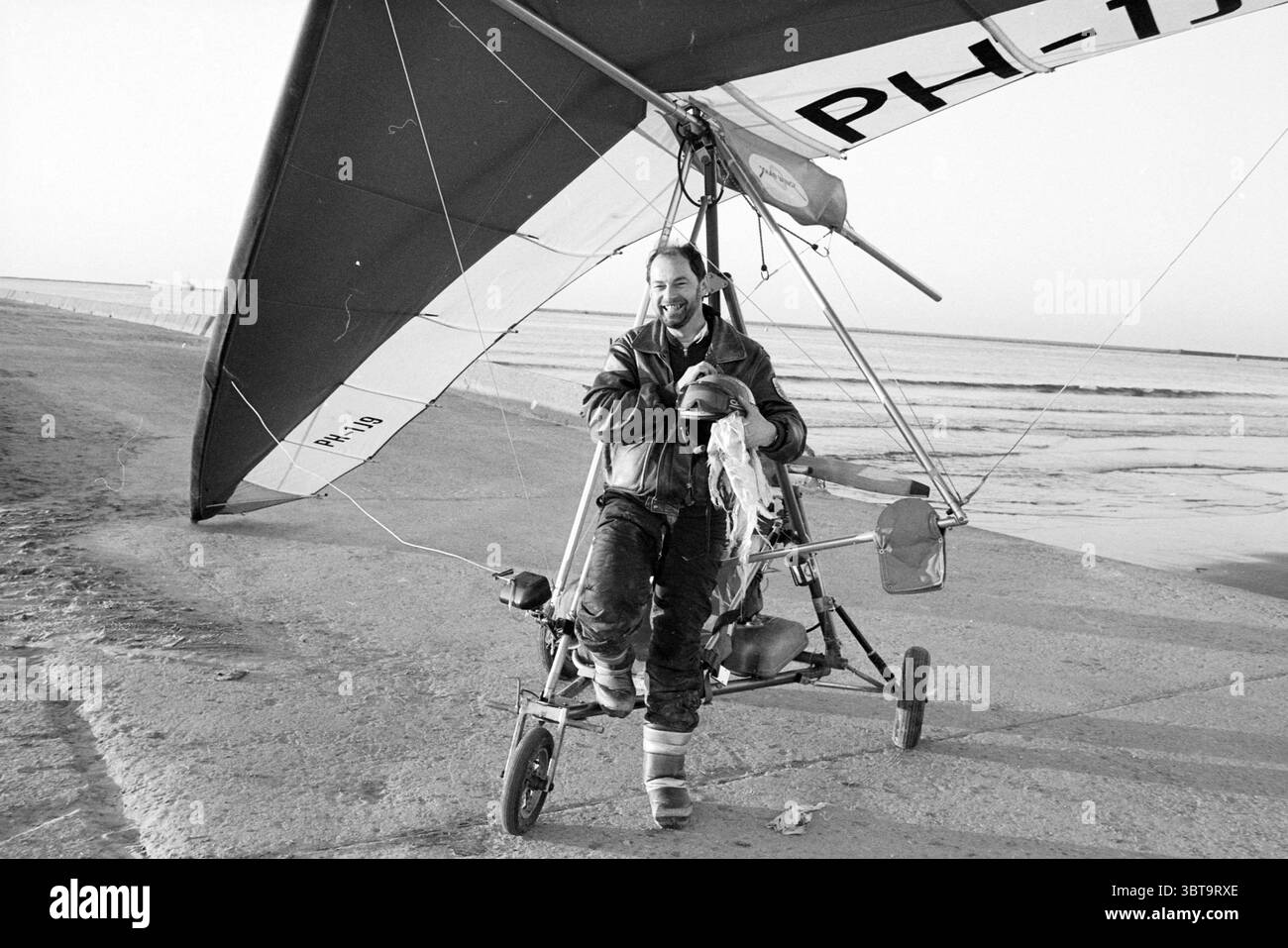Ed Lohman débarque d'urgence sur la plage. Cerfs-volants volants, Whizgle News, Dutch Desk, pays-Bas, 1950 - 2000 le 21-02-1989. Ces rubriques apparaissent dans l'image. La scène capture un homme debout à côté d'un petit avion léger, caractérisé par ses ailes de deltaplane distinctives. Les ailes, larges et étendues, projettent des ombres allongées sur le sol, mettant en valeur un design élégant qui évoque l'aventure aérienne. L'expression de l'homme transmet un sentiment de joie et d'accomplissement, avec un large sourire qui rayonne d'excitation. Il est habillé d'une veste de vol en cuir robuste qui suggère la chaleur Banque D'Images