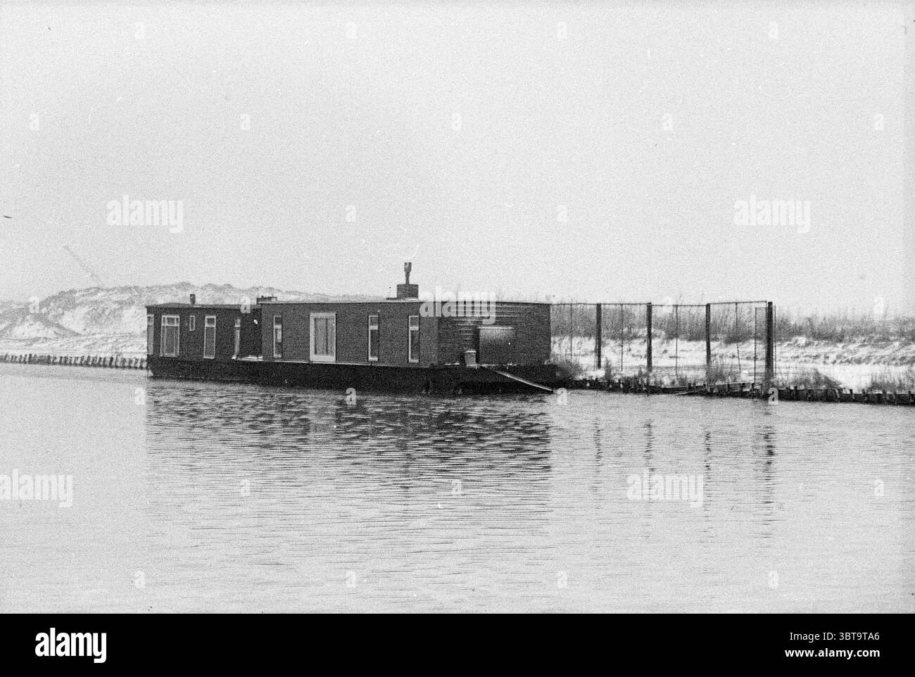 Amarrage pénal avec Houseboats, Whizgle News, Dutch Desk, pays-Bas, 1950 - 2000 le 06-12-1977. Ces rubriques sont illustrées dans l'image. Dans une voie navigable tranquille, une modeste péniche flotte sereinement, sa structure simpliste mais fonctionnelle. Le corps de la péniche est peint dans des teintes discrètes de brun foncé, suggérant une sensation de chaleur malgré l'environnement couvert. Sa forme rectangulaire est complétée par un toit plat, avec une cheminée qui fait allusion à un intérieur confortable où la chaleur pourrait rayonner contre le froid de la journée. Autour de la péniche est la surface vitreuse de l'eau Banque D'Images