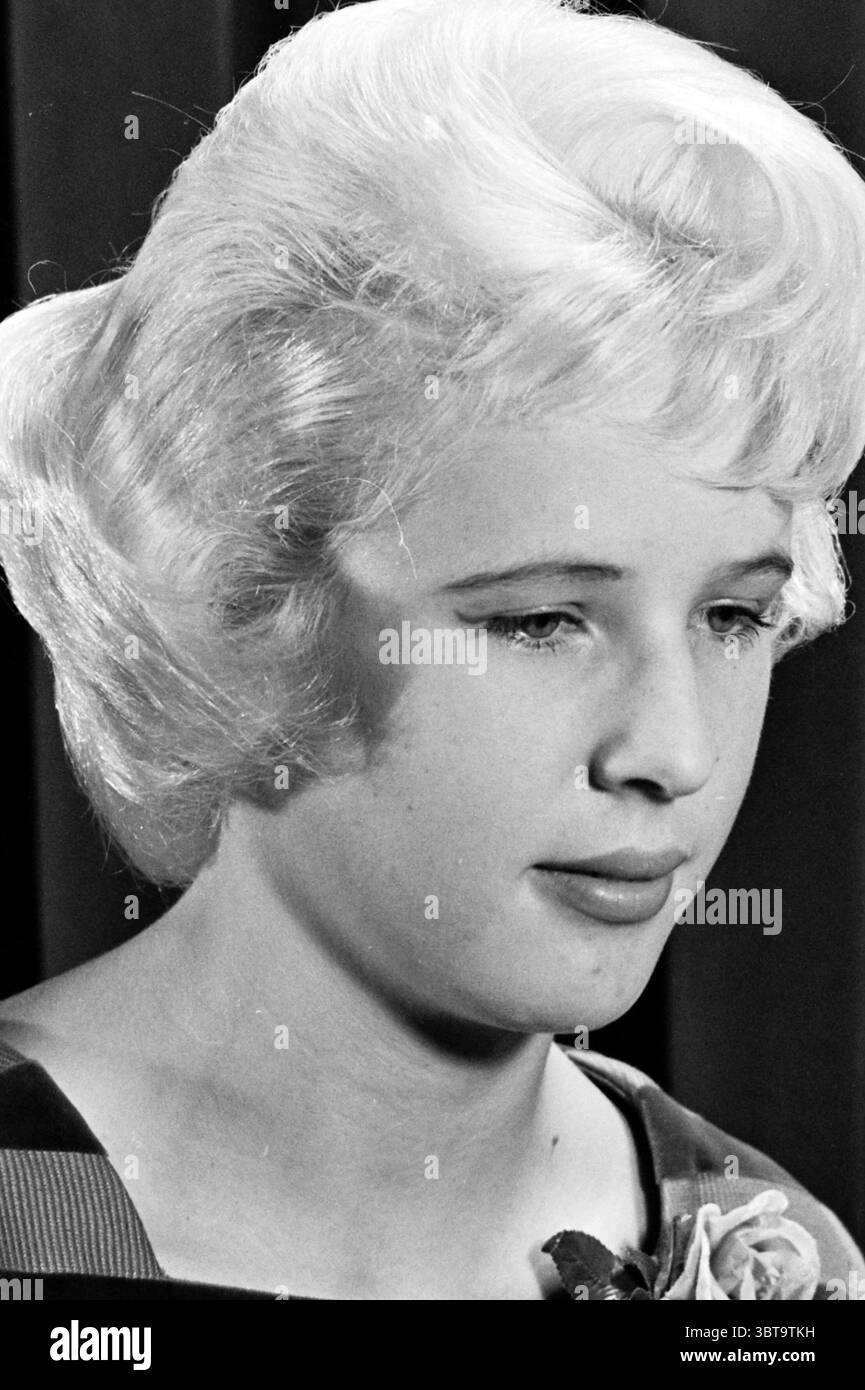 Coiffeurs Club Marcel coiffeurs et salons de coiffure, Whizgle News, Dutch Desk, pays-Bas, 1950 - 2000 le 25-09-1961. Ce sont les éléments de l'image. La scène met en scène une jeune femme avec une coiffure douce et volumineuse, caractérisée par des mèches lisses et ondulées qui encadrent son visage avec élégance. Ses cheveux, d’une teinte blond clair, contrastent subtilement sur un fond plus sombre, ce qui apporte profondeur et concentration à son expression. Elle a un regard doux et contemplatif, avec son regard dirigé légèrement vers le bas, créant une ambiance introspective. Sa peau semble claire et lisse, mettant en valeur le delica Banque D'Images