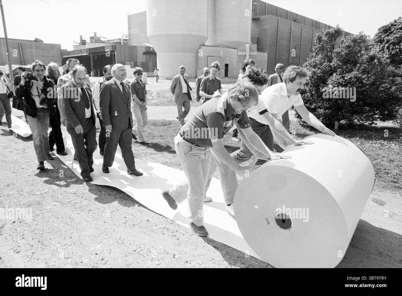 Election tour Den Uyl : IJmond Elections, Whizgle News, Dutch Desk, pays-Bas, 1950 - 2000 on 12-05-1981. L'image montre ces rubriques. La scène représente un groupe d'individus progressant le long d'une longue feuille de matériel qui traîne derrière eux. Ils traversent une surface extérieure accidentée, avec une toile de fond dominée par des structures industrielles, caractérisées par de grandes formes cylindriques et une série de bâtiments géométriques, suggérant un cadre manufacturier ou industriel. Les gens sont habillés dans un mélange de vêtements décontractés et semi-formels, indiquant un groupe diversifié avec divers rôles, l Banque D'Images