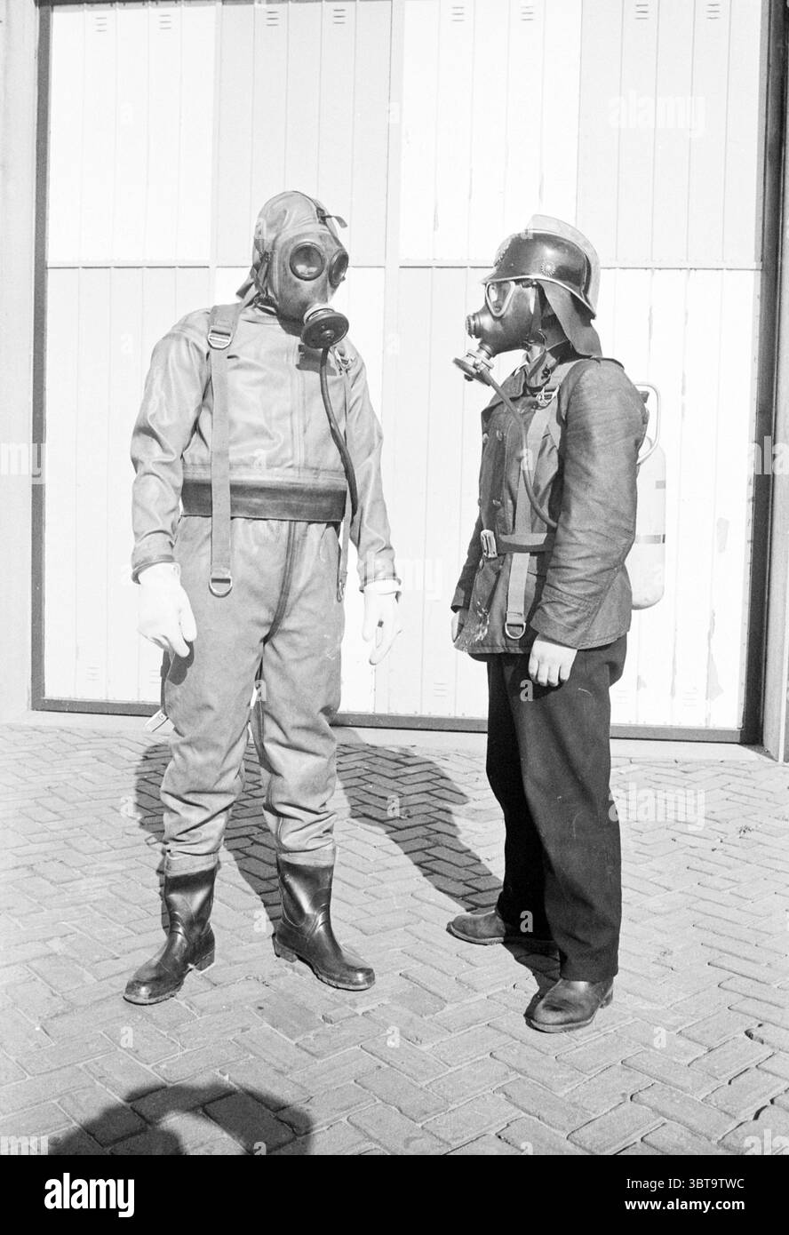 2 hommes avec masque à gaz et vêtements de protection., Whizgle News, Dutch Desk, pays-Bas, 1950 - 2000. Ce sont les éléments de l'image. La scène montre deux individus vêtus d'un équipement de protection distinct, suggérant un cadre lié à la sécurité ou à l'intervention d'urgence. Une personne est vêtue d'un costume sombre et robuste avec un masque à gaz, avec des gants et des bottes en caoutchouc, transmettant un sentiment d'urgence et de préparation aux conditions dangereuses. Le costume est probablement fait d'un matériau durable, et le masque à gaz obscurcit les traits du visage, laissant un sentiment d'anonymat. Le deuxième individu porte un sli Banque D'Images