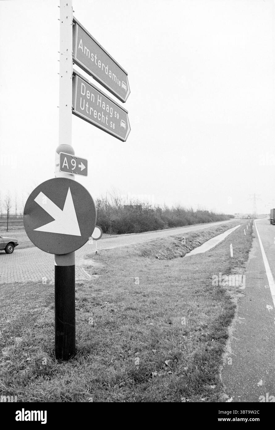 Wreck Road' Velsen au panneau ANWB faisant référence à A9 Velsen, Whizgle News, Dutch Desk, pays-Bas, 1950 - 2000 sur 26-11-1979. L'image montre ces rubriques. La scène présente un paysage sombre et monochrome, caractérisé par un ciel couvert d'humeur sombre qui donne une ambiance sombre. Un haut panneau de signalisation en métal se dresse bien en évidence sur le côté gauche, orné de plusieurs flèches directionnelles, chacune indiquant différentes destinations. Les panneaux sont peints dans une nuance de blanc, contrastant avec les tons plus sombres et discrets des environs. Sous le panneau, un grand panneau circulaire rouge avec un arro blanc et audacieux Banque D'Images