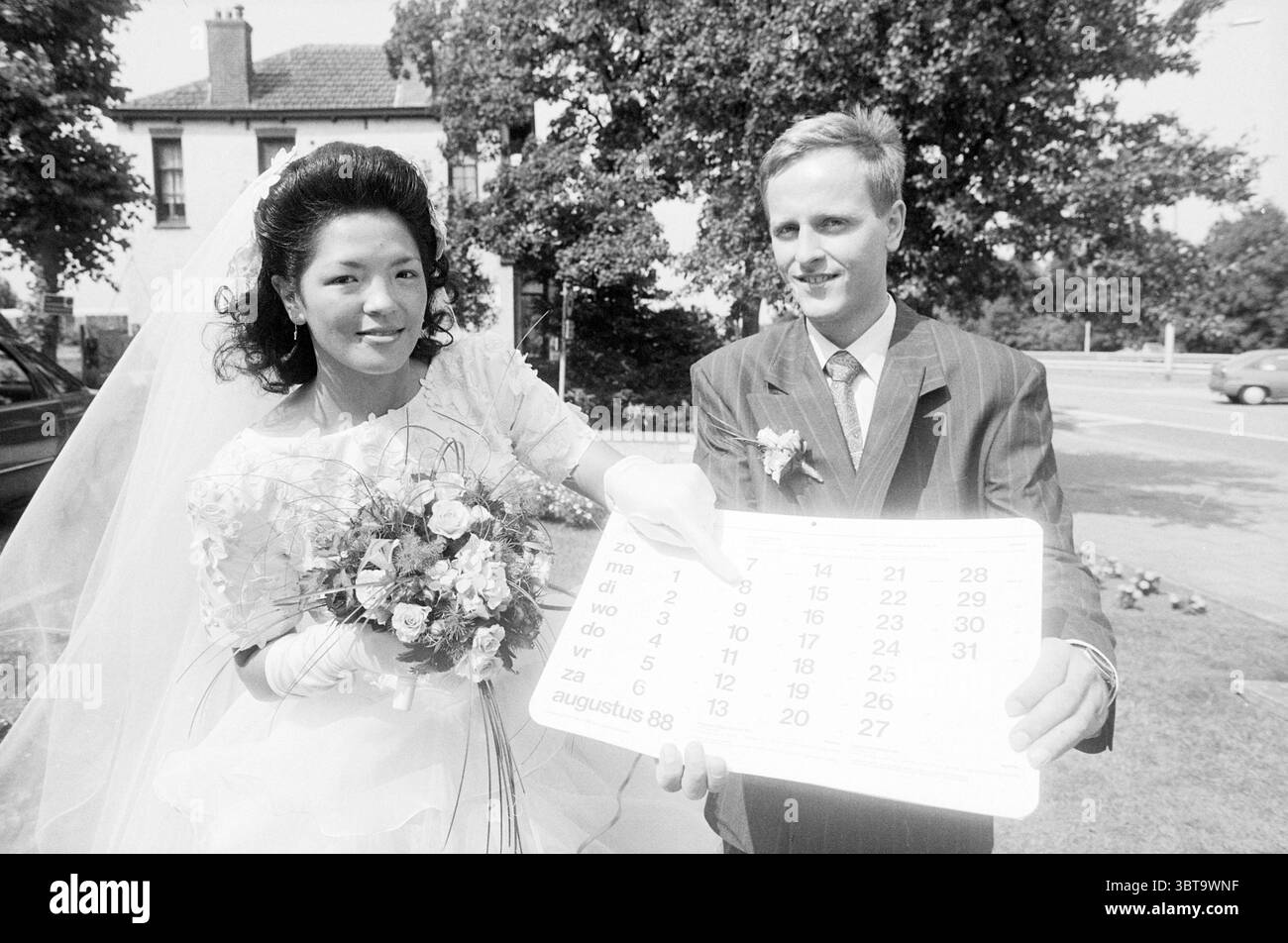 Mairie mariage Halfweg mariage Halfweg, Whizgle News, Dutch Desk, pays-Bas, 1950 - 2000 le 08-08-1988. Voici les rubriques de l'image. Dans un cadre extérieur ensoleillé, un couple se tient bien en vue au premier plan. La femme, vêtue d'une robe de mariée élégante, présente des détails de dentelle complexes et des manches bouffantes. Ses cheveux sont coiffés en vagues douces, ornés d'un voile délicat qui descend en cascade dans son dos. Elle tient un bouquet rempli de diverses fleurs, mettant en valeur un mélange de teintes douces qui contrastent délicieusement avec sa robe. À côté d'elle, un homme dans un costume bien taillé se confie Banque D'Images