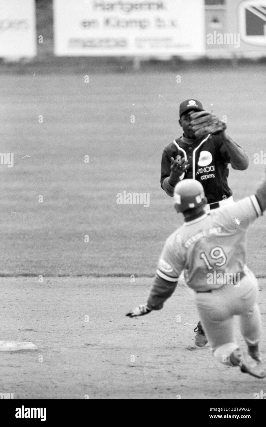 Baseball : Pioniers - Sparta, Whizgle News, Dutch Desk, pays-Bas, 1950 - 2000 on 15-05-1996. L'image montre ces rubriques. La scène capture un moment dynamique sur un diamant de baseball. D'un côté, un joueur, vêtu d'un uniforme sombre avec une casquette remarquable, est accroupi, prêt et alerte. Son attention est dirigée vers un coureur approchant du premier plan, qui porte un maillot de couleur claire orné du numéro 19. Le coureur est à mi-foulée, transmettant un sentiment d'urgence et d'action alors que ses pointes creusent dans la terre. Le fond est un mélange de vert herbeux et de bruns terreux, typique Banque D'Images