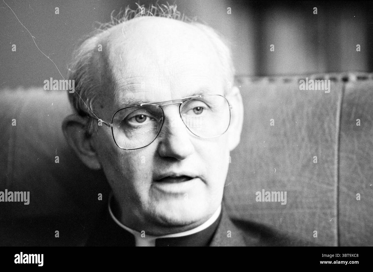 Mgr. Bomers Haarlem Roman Catholic Church Haarlem pays-Bas, Whizgle News, Dutch Desk, pays-Bas, 1950 - 2000 on 10-05-1987. L'image contient ces rubriques. La scène met en scène un homme âgé avec une expression réfléchie, assis dans un espace intérieur confortable. Il a les cheveux blancs amincis et porte des lunettes à bordure métallique qui soulignent ses yeux brillants. Sa tenue vestimentaire se compose d'un col clérical sombre, suggérant un contexte religieux. Le cadre est chaleureusement éclairé, avec une lumière douce et naturelle filtrant à travers une fenêtre voisine, créant des ombres douces qui mettent en valeur les contours de son visage. Le backgro Banque D'Images