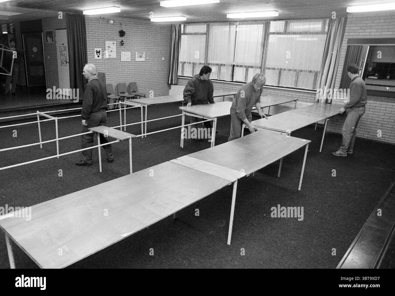 Évacuation du tournoi d'échecs des hauts fourneaux en raison d'une fuite HST Wijk aan Zee, Whizgle News, Dutch Desk, pays-Bas, 1950 - 2000 on 27-01-1990. Ces rubriques sont illustrées dans l'image. La scène capture un espace intérieur spacieux, probablement une salle communautaire ou une salle polyvalente, caractérisé par son design simple et fonctionnel. Le sol est recouvert de moquette sombre, qui contraste avec les murs texturés de couleur claire. Ces murs ont quelques affiches ou avis épinglés, ajoutant une touche de couleur et d'activité à l'environnement autrement tamisé. Quatre personnes sont activement engagées dans Moving et arra Banque D'Images
