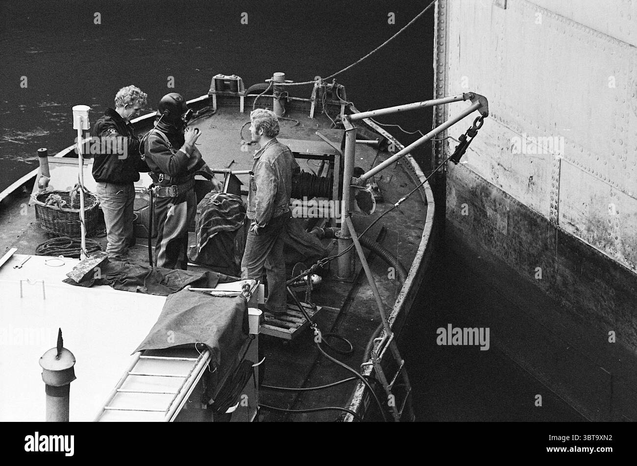 Travailler à l'écluse, Whizgle News, Dutch Desk, pays-Bas, 1950 - 2000 sur 13-10-1969. L'image inclut ces rubriques. La scène représente un petit bateau amarré à côté d'une grande structure, probablement un entrepôt ou un navire. Trois individus sont situés sur le pont du bateau, engagés dans la conversation. Une personne se tient à gauche, vêtue d'une veste sombre, et semble concentrée sur l'interaction avec les autres. Le personnage au centre porte une combinaison humide, suggérant qu'ils peuvent être impliqués dans des travaux sous-marins ou de la plongée. Le troisième individu, positionné à droite, a les cheveux plus clairs et porte un d Banque D'Images