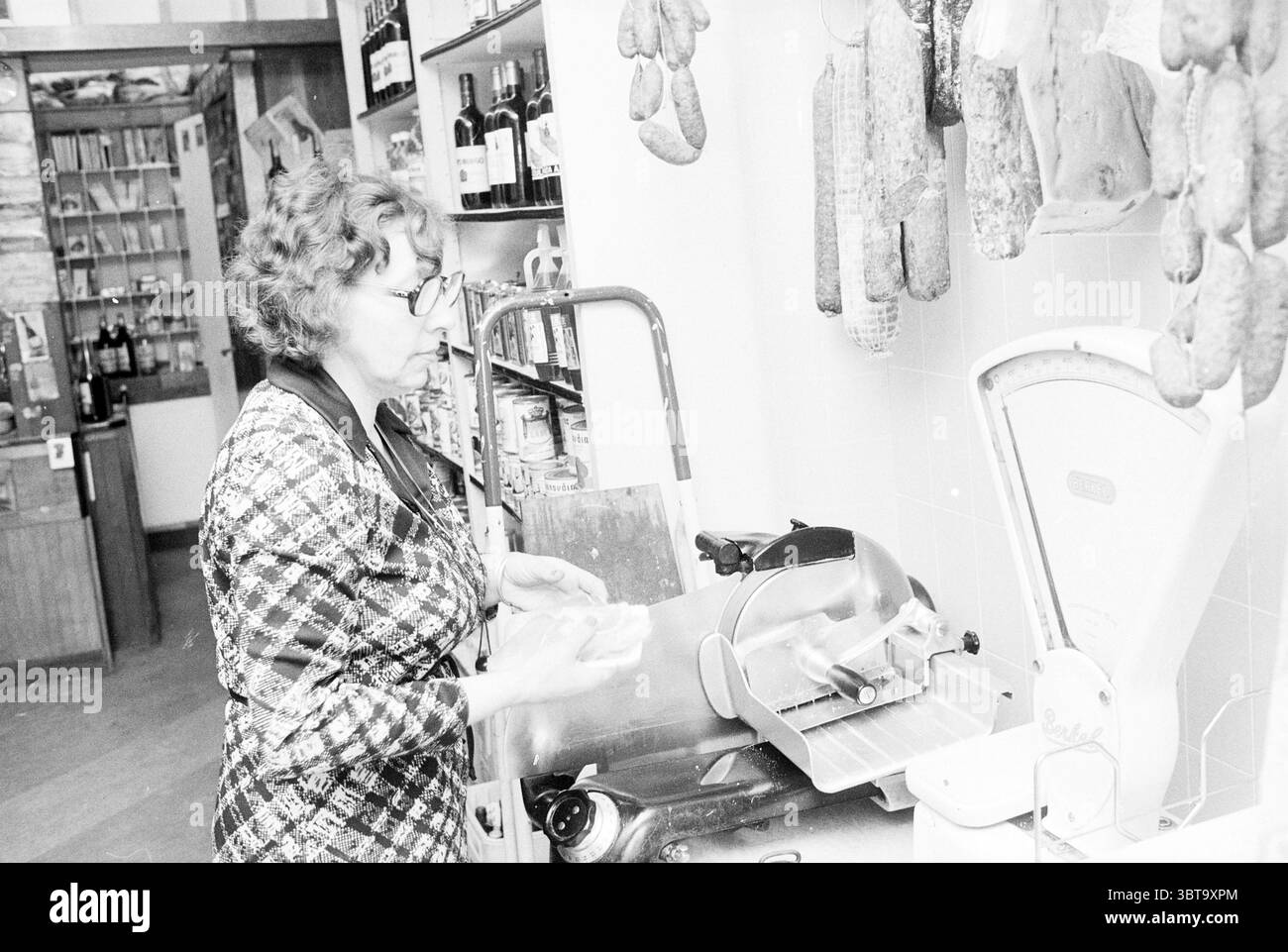 The Spanish Shop, Whizgle News, Dutch Desk, pays-Bas, 1950 - 2000 en 1977. L'image montre ces rubriques. La scène révèle une charcuterie ou une boucherie confortable de style vintage. À gauche, les murs sont ornés d'un éventail de viandes salées suspendues, certaines enveloppées de tissu, mettant en valeur un charme rustique. Leurs teintes brun doré et rougeâtres créent une atmosphère chaleureuse, contrastant avec les carreaux blancs propres et lumineux du mur. Au premier plan, une femme se tient sur un comptoir, son expression focalisée révélant son dévouement à son métier. Elle a les cheveux bouclés, coiffée soigneusement, et porte des lunettes qui lui prêtent un sch Banque D'Images