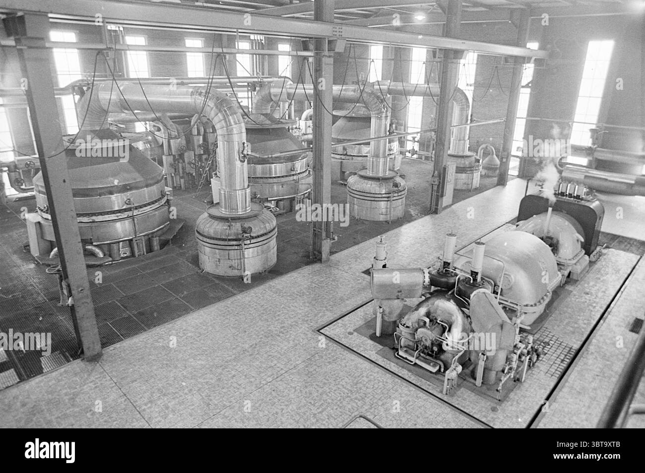 Usine d'acide nitrique Hoogovens Mekog. Hoogovens, Whizgle News, Dutch Desk, pays-Bas, 1950 - 2000 sur 10-07-1974. L'image inclut ces rubriques. La scène présente un grand espace industriel rempli de machines complexes. Plusieurs grands vaisseaux arrondis dominent le premier plan, leurs surfaces métalliques brillant doucement sous les plafonniers. Ces récipients, éventuellement en acier inoxydable, sont reliés par un réseau complexe de tuyaux qui sillonnent la pièce, réfléchissant la lumière ambiante. Le décor présente de hauts plafonds soutenus par d'épaisses poutres métalliques, créant un sentiment de grandeur Banque D'Images