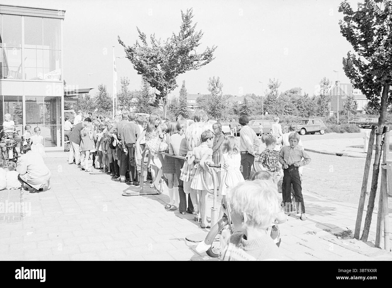 1st Customers Stuif Stuif Stuif Stuif Stuif, Whizgle News, Dutch Desk, pays-Bas, 1950 - 2000 le 14-07-1969. Voici les rubriques de l'image. La scène capture un cadre extérieur animé, caractérisé par une longue file de personnes qui attendent leur tour. À gauche, un groupe est rassemblé étroitement, avec quelques individus assis sur le sol, peut-être au repos tandis que d'autres se tiennent debout. Les personnes en ligne sont principalement des enfants et des adolescents, habillés dans une variété de styles vestimentaires qui reflètent une esthétique rétro. Leur tenue vestimentaire comprend des robes à motifs, des chemises rayées et des vestes légères aux couleurs pastel Banque D'Images