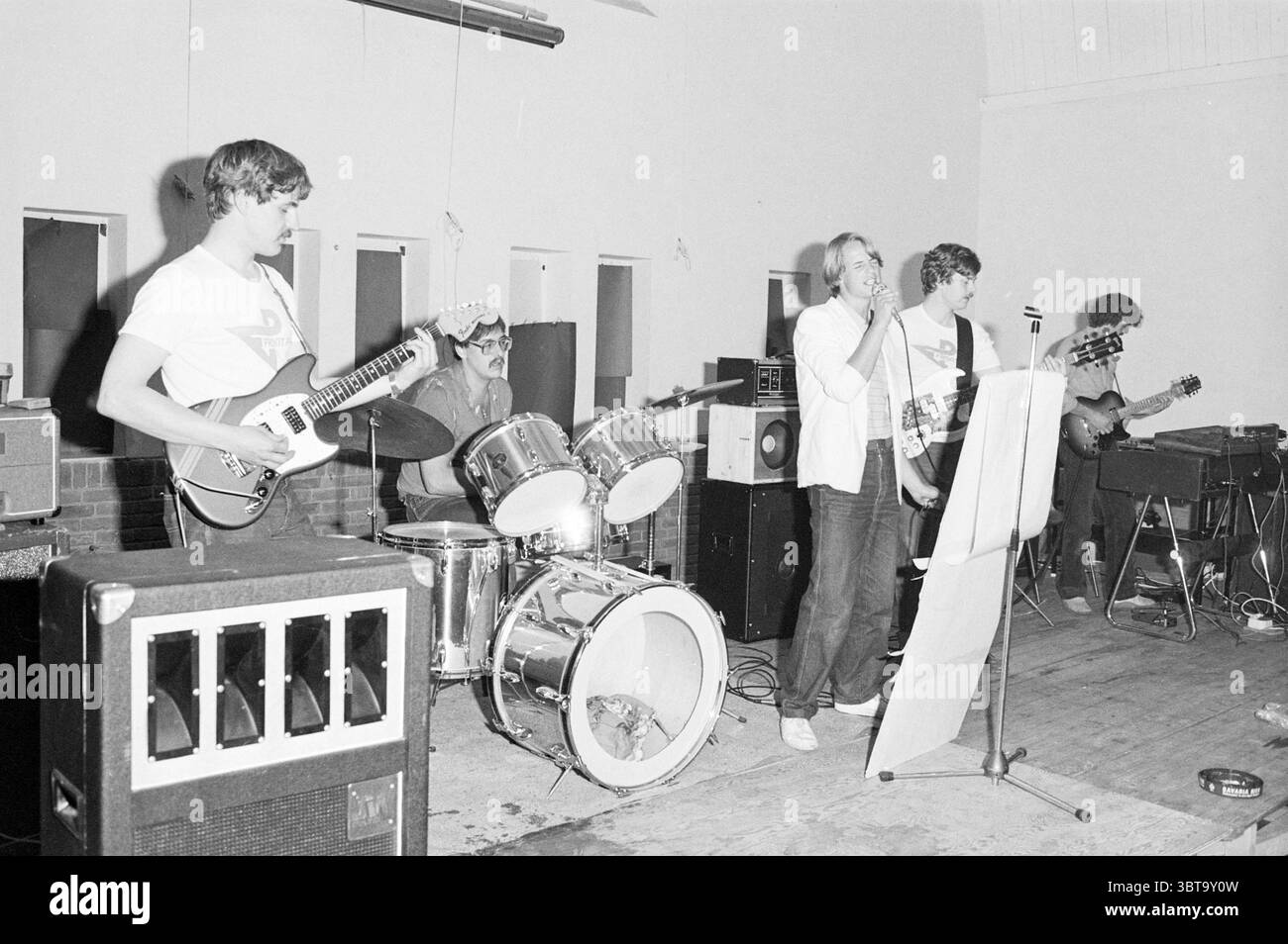 Performance 'Front page' dans Velsen-n' Music Velsen-Noord, Whizgle News, Dutch Desk, pays-Bas, 1950 - 2000 sur 29-05-1982. Voici les rubriques de l'image. La scène met en scène un groupe animé se produisant sur une scène simple dans un lieu intérieur, qui a une atmosphère rustique et décontractée. La toile de fond présente un mur sans ornement, et le sol semble être en bois, suggérant un espace accueillant et accueillant. Au premier plan, un chanteur masculin se tient au centre, saisissant un microphone, s'engageant activement avec la foule. Il porte une chemise décontractée et a une expression confiante, rayonnant d'énergie. À Banque D'Images