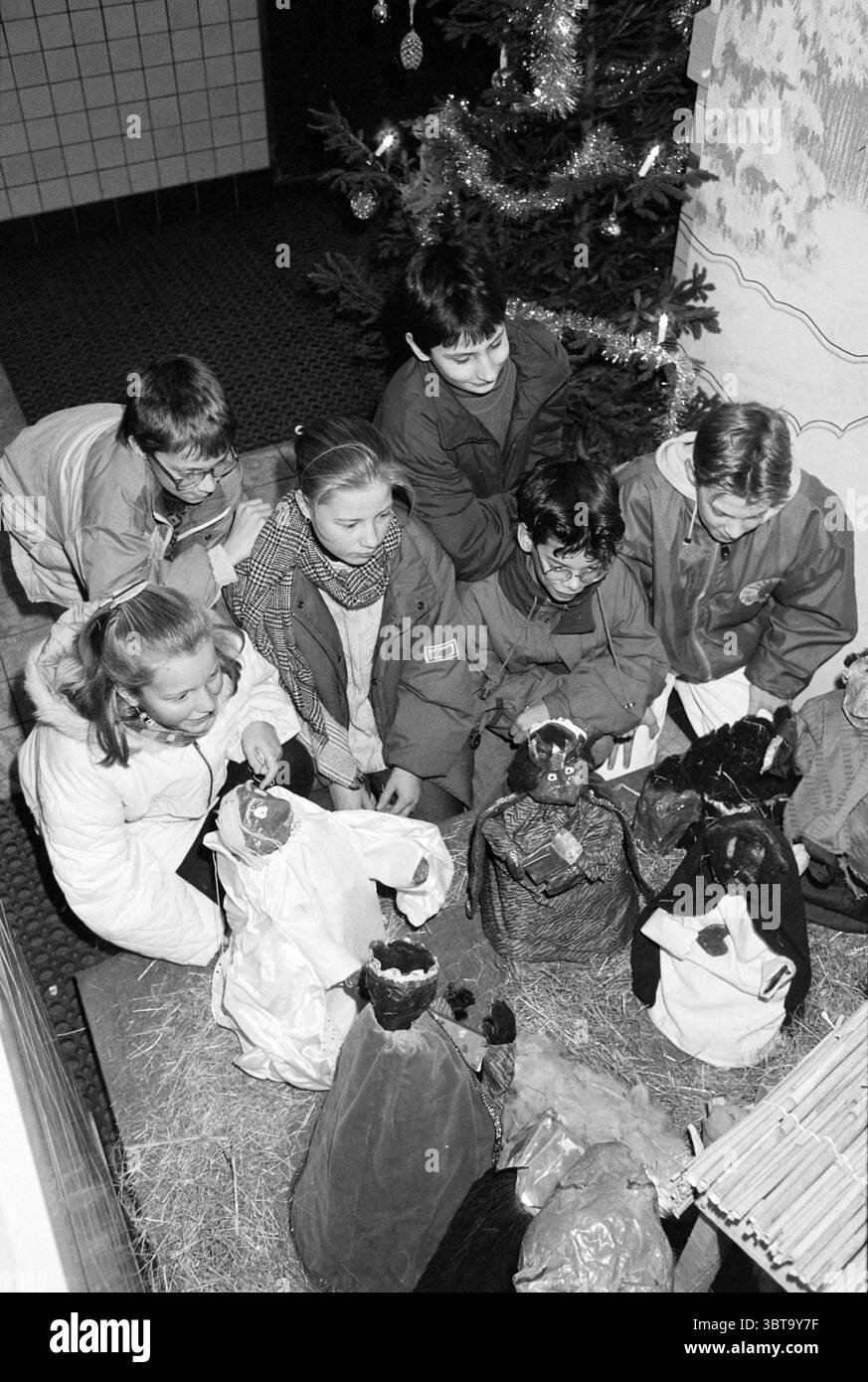Fierté de Noël Cardinal Alfrink School, Whizgle News, Dutch Desk, pays-Bas, 1950 - 2000 on 16-12-1992. L'image contient ces rubriques. Dans un cadre intérieur confortable, un groupe de six enfants se réunit étroitement autour d'une scène de nativité avec des figurines artisanales. La toile de fond est ornée d'un sapin de Noël clairsemé et décoré qui ajoute une touche festive, avec des ornements scintillants contrastant avec l'ambiance sombre de l'espace. Les expressions des enfants reflètent la curiosité et l'admiration comme ils observent les figures, chacune soigneusement habillée de différentes couleurs de tissu. Les couleurs dominantes dans le sce Banque D'Images