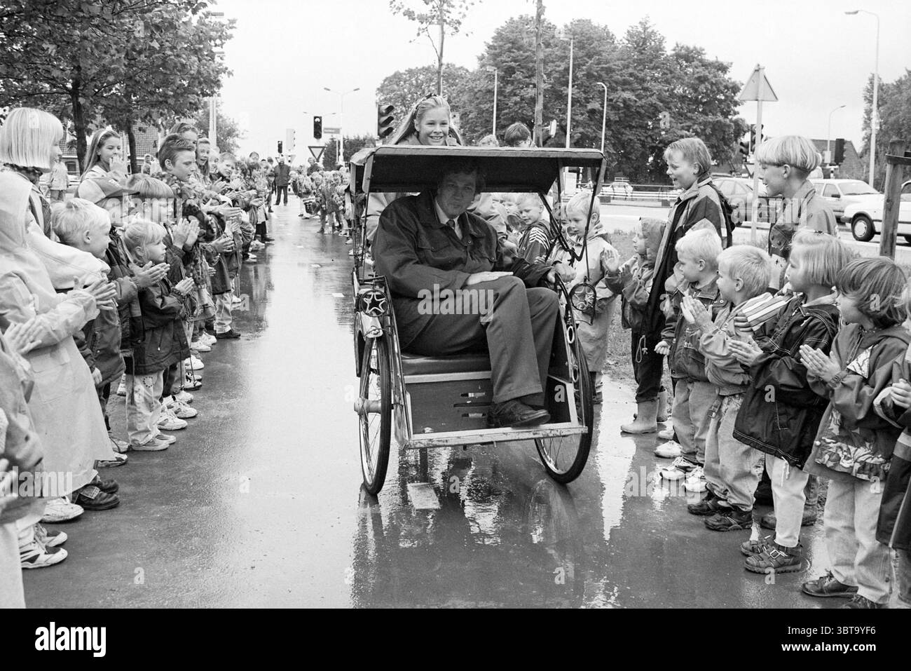 Ouverture de la piste cyclable de Noorderdreef Nieuw-Vennep Nieuw-Vennep Noorderdreef, Whizgle News, Dutch Desk, pays-Bas, 1950 - 2000 le 15-06-1995. Ces rubriques apparaissent dans l'image. Dans un cadre urbain animé, une scène animée se déroule dans une rue baignée de pluie, scintillant sous le ciel gris tamisé. Des gens de différents âges bordent le trottoir, créant une tapisserie vibrante d'excitation. Les enfants, principalement en imperméables, manifestent des expressions de joie et d’anticipation, leurs visages illuminés d’enthousiasme. Ils applaudissent et applaudissent, créant une atmosphère de fête. Au centre de la scène, un Banque D'Images