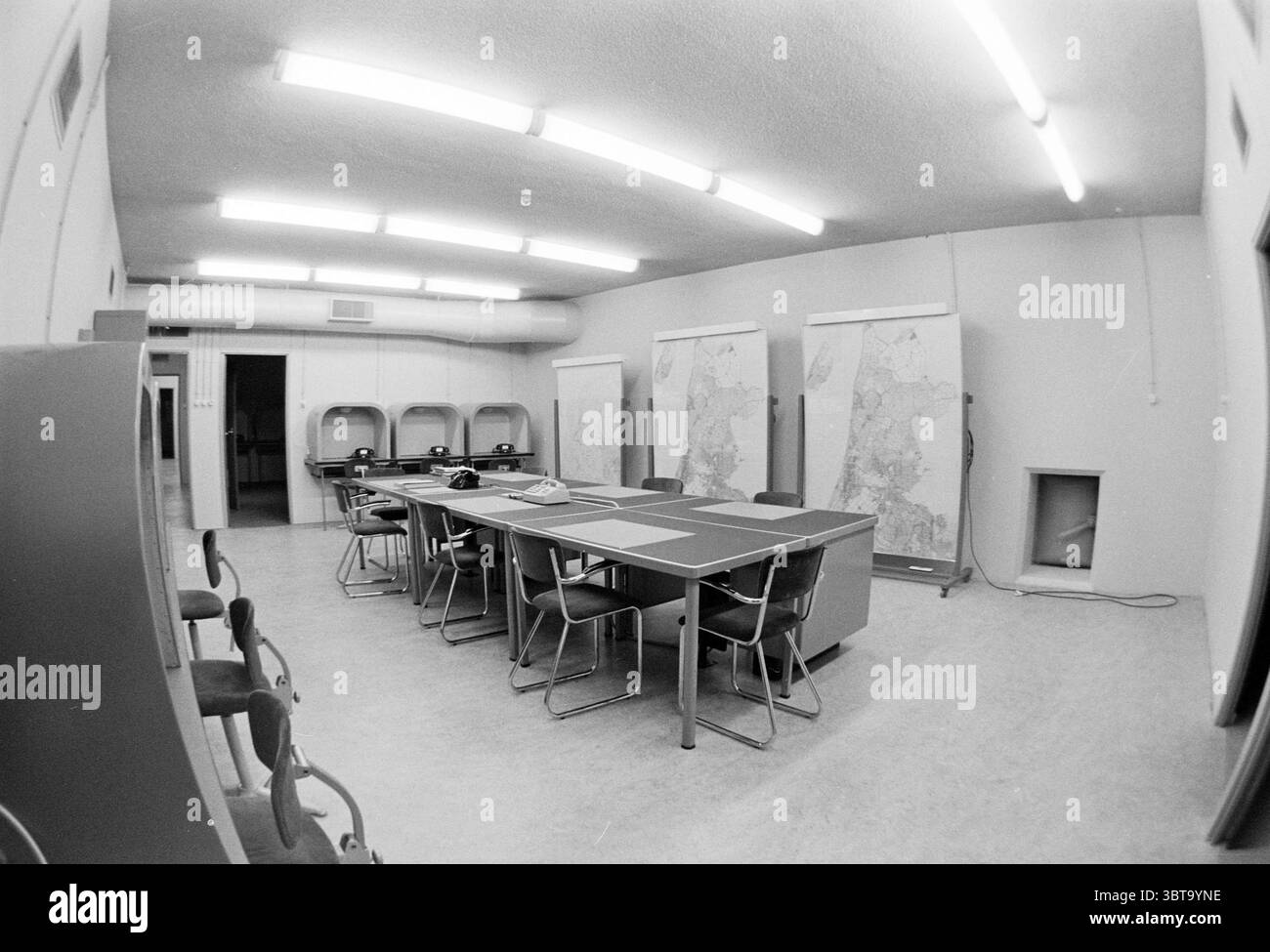 Intérieur d'un bureau., Whizgle News, Dutch Desk, pays-Bas, 1950 - 2000. Ce sont les éléments de l'image. La scène représente une salle spacieuse, quelque peu stérile, conçue pour des réunions ou des discussions. Les murs sont peints d'une couleur claire et neutre, contribuant à une ambiance minimaliste. Une longue table rectangulaire domine le centre, entourée d'une série de chaises à ossature métallique. Les chaises semblent fonctionnelles mais manquent d'une touche de confort, ce qui suggère de mettre l'accent sur l'efficacité plutôt que sur la détente. Au-dessus, des lumières fluorescentes brillantes projettent un éclairage net, améliorant la sensation clinique de la spac Banque D'Images