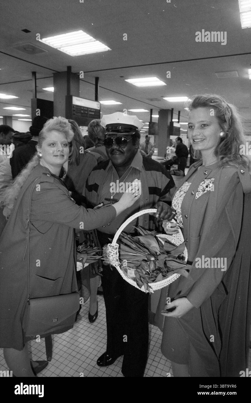 Fats Domino Arrival Schiphol Schiphol, Whizgle News, Dutch Desk, pays-Bas, 1950 - 2000 le 09-04-1991. L'image montre ces rubriques. La scène représente un environnement intérieur animé, probablement un aéroport ou un terminal public, rempli de diverses personnes engagées dans diverses activités. La composition comporte trois figures importantes occupant le premier plan. Deux femmes se tiennent de chaque côté d'un homme. Une femme a des cheveux volumineux et de couleur claire, portant une veste sombre qui contraste avec son teint clair. Son expression semble amicale et engagée alors qu'elle interagit avec l'homme. Le se Banque D'Images