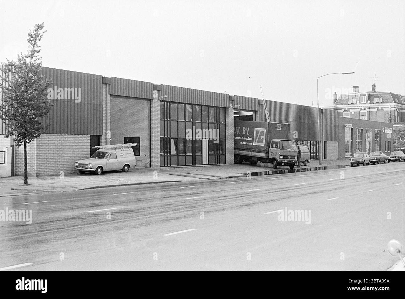 Outdoor Timber trade v.d. Belt Spaarndamseweg bois extérieur Haarlem Spaarndamseweg pays-Bas, Whizgle News, Dutch Desk, pays-Bas, 1950 - 2000 on 28-09-1976. L'image montre ces rubriques. La scène représente l'extérieur d'un bâtiment commercial sur un ciel gris et couvert, suggérant une atmosphère sombre et silencieuse. La structure présente une combinaison de brique et de métal, avec une grande fenêtre en verre qui reflète la lumière terne. Le design du bâtiment est fonctionnel et moderne, caractérisé par des lignes épurées et une orientation horizontale. Au premier plan, il y a une large route asphaltée, slig Banque D'Images