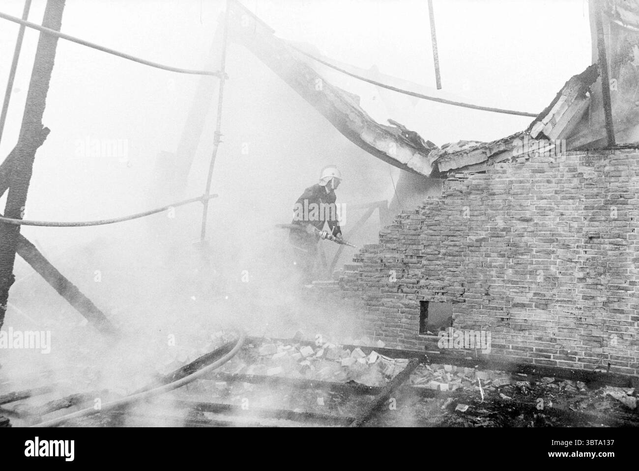 Hangar à ampoules à feu fa. Willemsen Hillegom Fire brigade Hillegom, Whizgle News, Dutch Desk, pays-Bas, 1950 - 2000 on 02-03-1980. L'image montre ces rubriques. La scène capture un moment sombre et intense au milieu des suites d'un incendie. La fumée circule partout, créant une atmosphère épaisse et brumeuse qui obscurcit la visibilité. Au premier plan, un pompier, équipé d'un équipement de protection, se tient en évidence. La figure est partiellement éclairée, contrastant avec le fond plus sombre et plus chaotique. Leur expression est ciblée et déterminée, soulignant le courage requis dans un tel désastre Banque D'Images
