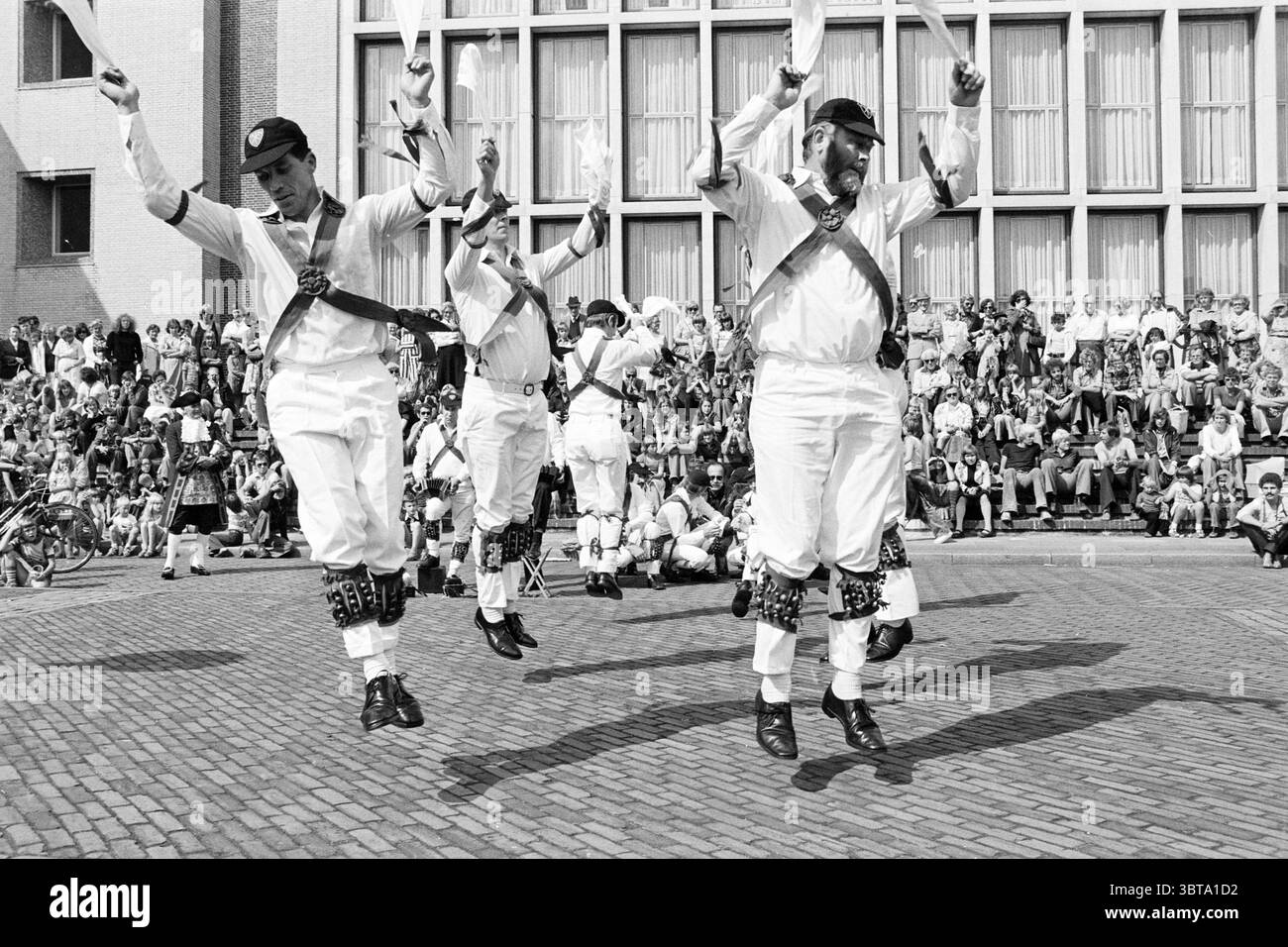 Morris danseurs à IJm. Danse, Whizgle News, Dutch Desk, pays-Bas, 1950 - 2000 le 03-09-1977. L'image contient ces rubriques. Dans la scène, un groupe de cinq hommes est engagé dans une danse traditionnelle animée, leurs mouvements énergiques et synchronisés. Vêtus de pantalons et chemises blancs, ils portent des gilets noirs ornés de ceintures décoratives qui créent un contraste saisissant avec leurs tenues. Chaque danseur arbore une casquette noire, ajoutant à l'uniformité de leur apparence. Les danseurs sont capturés à mi-saut, les bras levés, créant une impression de joie et de célébration. Le Mo Banque D'Images