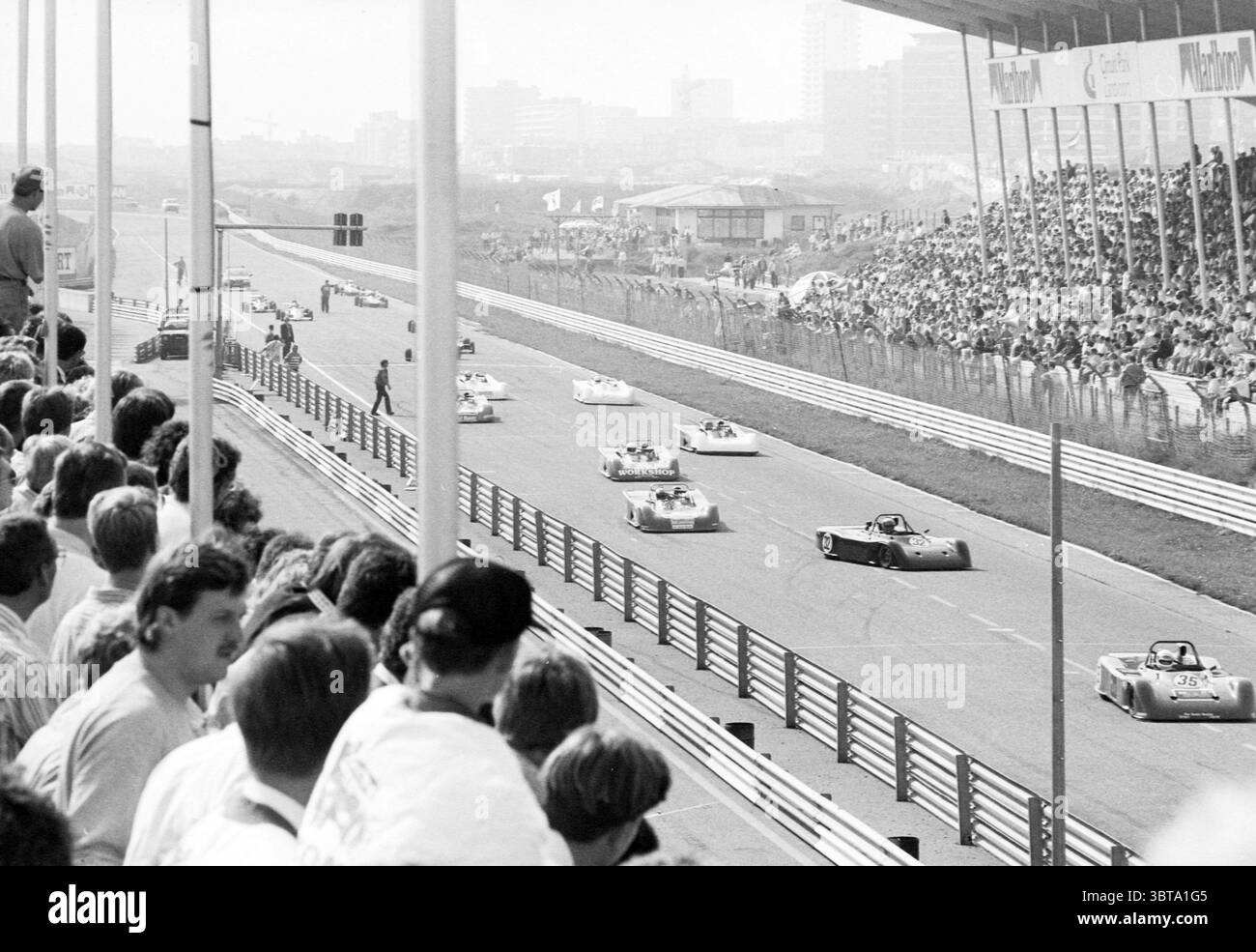 Circuit automobile Zandvoort Zandvoort, Whizgle News, Dutch Desk, pays-Bas, 1950 - 2000 en 1989. Ces rubriques apparaissent dans l'image. La scène visuelle capture un événement de course dynamique se déroulant sur un large circuit, où plusieurs petites voitures de course sont en mouvement. Les voitures, principalement dans des tons de blanc et de couleurs claires, sont élégantes et basses au sol, mettant l'accent sur la vitesse et l'agilité. Les voitures de course sont disposées en formation, suggérant une compétition passionnante alors qu'elles manœuvrent le long de la piste. Au premier plan, une foule de spectateurs peut être vue assis dans les gradins, leur attention concentrée sur t Banque D'Images