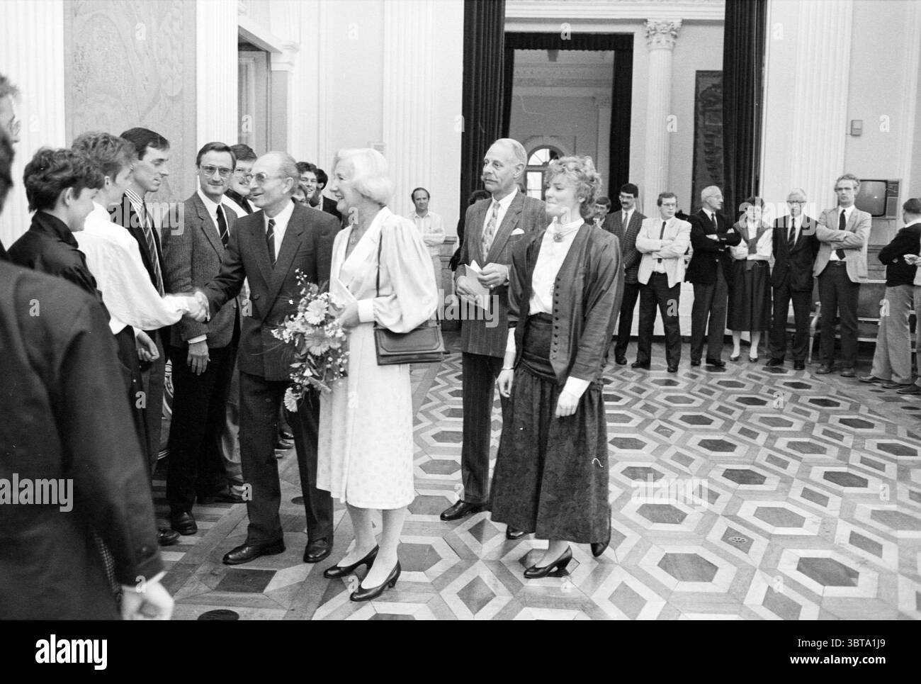 Pourrait. Prix Mr. de Korte. Royal Honors, Whizgle News, Dutch Desk, pays-Bas, 1950 - 2000 le 13-11-1987. L'image contient ces rubriques. La scène se déroule dans un intérieur spacieux et élégamment conçu, caractérisé par de hauts plafonds et des planchers en bois poli dans un motif géométrique. La lumière jaillit de grandes fenêtres, projetant des ombres douces qui rehaussent l'atmosphère lumineuse et accueillante. Un groupe de personnes se rassemble au premier plan, vêtu d'une tenue formelle. Leurs vêtements présentent un mélange de styles : certains portent des costumes et des cravates, tandis que d'autres sont dans des robes, ajoutant de la variété à la scène. L'ensem Banque D'Images