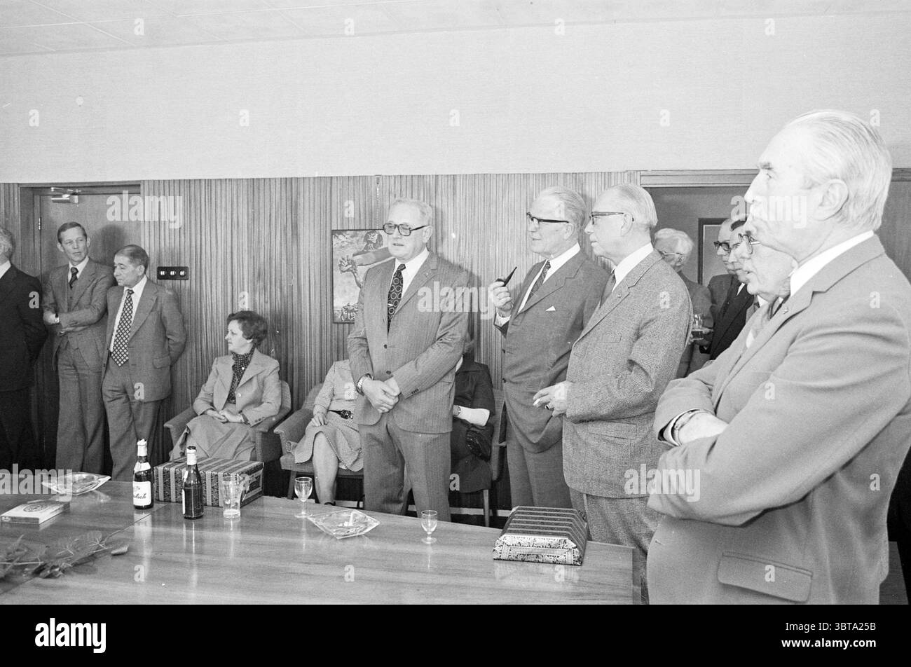 Les examinateurs CBR disent adieu Rijswijk C.B.R., Whizgle News, Dutch Desk, pays-Bas, 1950 - 2000 le 20-03-1976. Ce sont les éléments de l'image. La scène se déroule dans un cadre intérieur formel, probablement une salle de conférence ou un espace de rassemblement similaire, caractérisé par des panneaux en bois qui ajoutent une touche de chaleur. Une longue table s’étend au premier plan, ornée d’un arrangement de petits verres et de quelques bouteilles de boissons, suggérant une atmosphère raffinée. Au milieu du terrain, un groupe de plusieurs individus se tient en demi-cercle, chacun habillé dans des costumes élégants et sur mesure, indicatifs d'un pr Banque D'Images