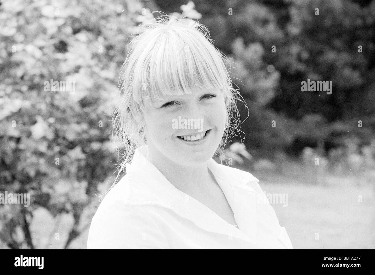 Rire du jour ................... Bokkedoorns Persons, Whizgle News, Dutch Desk, pays-Bas, 1950 - 2000 on 27-07-1978. L'image contient ces rubriques. La scène capture une jeune femme debout à l'extérieur, dégageant une présence chaleureuse et invitante. Elle a les cheveux blonds clairs coiffés de franges douces et de couches à la longueur des épaules, créant un cadre doux autour de son visage. Son sourire est brillant et authentique, suggérant un comportement joyeux, avec ses yeux scintillants dans un moment de joie. Autour d'elle est un environnement luxuriant, avec de la verdure qui fait allusion à un cadre naturel. L'arrière-plan est légèrement Banque D'Images