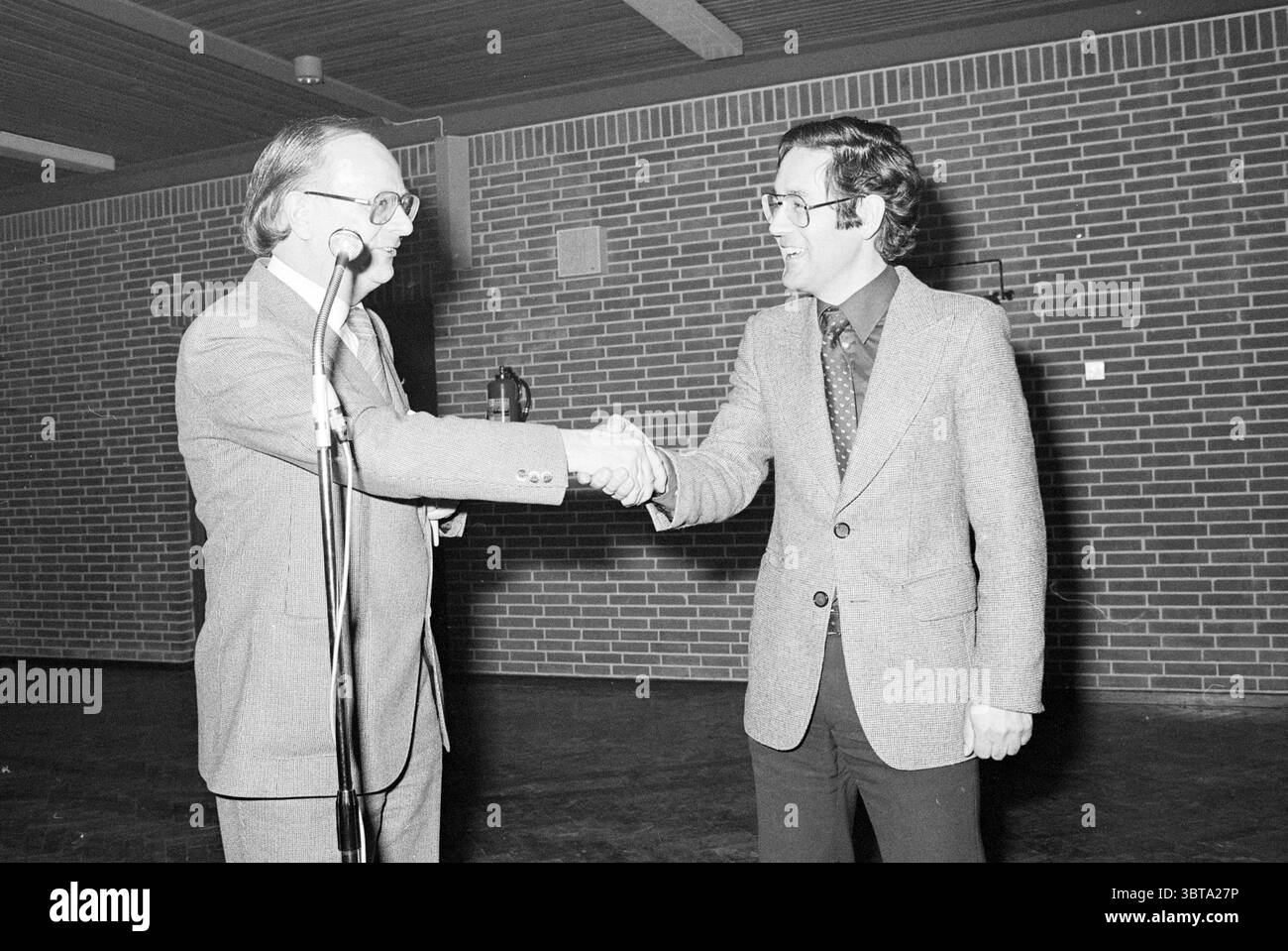Concours de remise de prix cérémonie de remise de prix Heemskerk Heemskerk, Whizgle News, Dutch Desk, pays-Bas, 1950 - 2000 le 07-05-1979. Voici les rubriques de l'image. Dans la scène, deux hommes sont engagés dans une poignée de main, transmettant un moment d'accord ou de camaraderie. Un homme est habillé d'un costume avec une cravate, avec un motif subtil, tandis que l'autre arbore une chemise à motifs plus décontractée sous un blazer, suggérant une atmosphère amicale. Le cadre est un espace intérieur au design épuré et minimaliste, caractérisé par des murs de briques texturés qui créent une toile de fond chaleureuse et terreuse. Le Lightin Banque D'Images