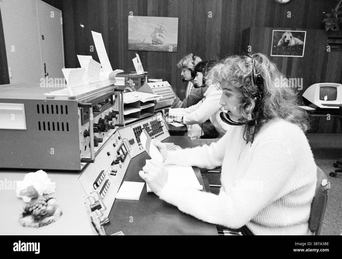 Scheveningen radios IJmuiden Radio, Whizgle News, Dutch Desk, pays-Bas, 1950 - 2000 on 28-12-1981. L'image montre ces rubriques. La scène dispose d'une salle remplie de technologie et d'équipements rétro, sur fond de boiseries sombres. Deux femmes sont engagées dans leur travail, assises à une console ornée d'un ensemble de boutons, cadrans et leviers. Les deux portent des casques, indiquant leur rôle dans la communication ou la coordination. Une femme, positionnée au premier plan, a des cheveux bouclés volumineux et se concentre sur la rédaction de notes ou la gestion de la paperasse, son expression un mélange de concentr Banque D'Images