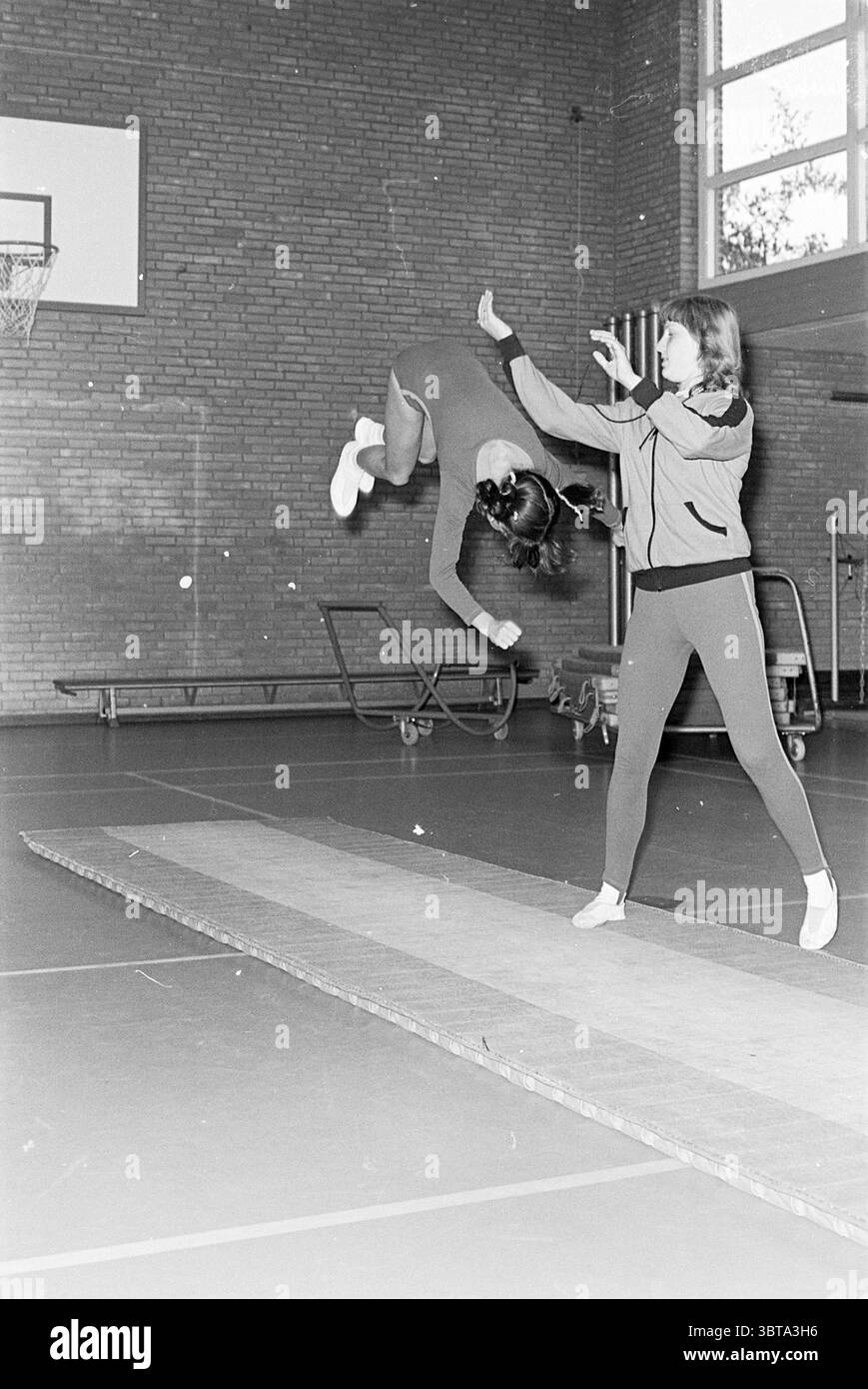 Gymnastes avec professeur de gymnastique Waterloohal Driehuis Gymnastics, Whizgle News, Dutch Desk, pays-Bas, 1950 - 2000 le 22-10-1975. Voici les rubriques de l'image. Dans un gymnase spacieux caractérisé par de hauts plafonds et des murs de briques apparentes, la scène met en scène deux individus engagés dans une activité physique. Une personne est en plein air, effectuant un flip dynamique, montrant un niveau impressionnant de compétence et d'agilité. Cet athlète porte une tenue ajustée qui met l'accent sur le mouvement, probablement dans des tons de rouge, ce qui lui permet de se démarquer sur la toile de fond discrète. Leurs cheveux coulent derrière eux, ajoutant à Banque D'Images