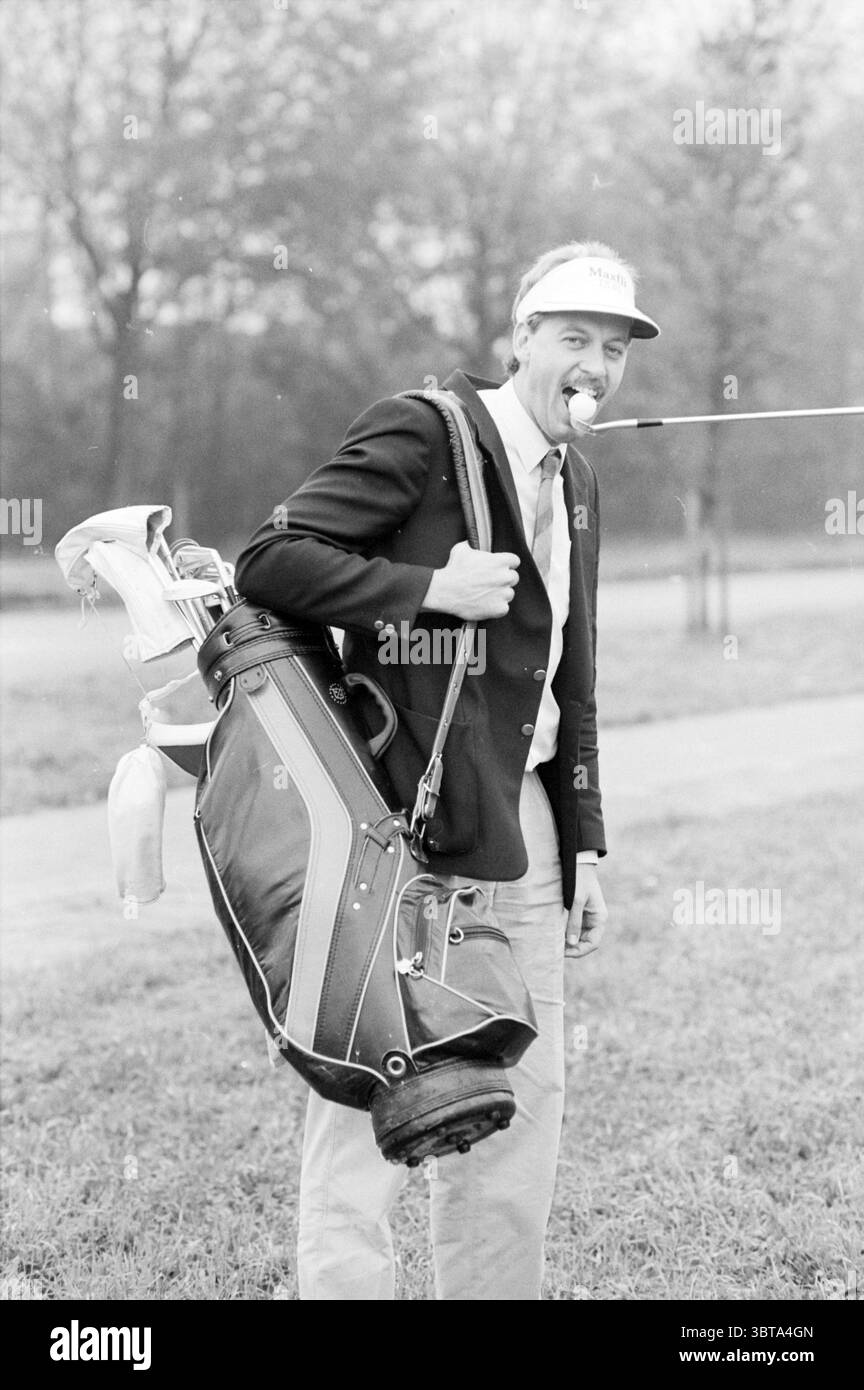 Golfer Hits ball., Whizgle News, Dutch Desk, pays-Bas, 1950 - 2000 sur 10-1988. L'image inclut ces rubriques. La scène capture un jeune homme debout sur une zone herbeuse qui semble faire partie d'un terrain de golf. Il est habillé d'un blazer sur mesure et d'un pantalon de couleur claire, suggérant une approche chic-décontractée ou formelle du golf. Un sac de golf, rempli de clubs, est accroché sur son épaule, et il tient un club de golf dans sa bouche de manière ludique, mettant en valeur un comportement léger. L'homme a les cheveux courts et une casquette qui ajoute une touche sportive à sa tenue. L'arrière-plan présente un paysage flou de Banque D'Images