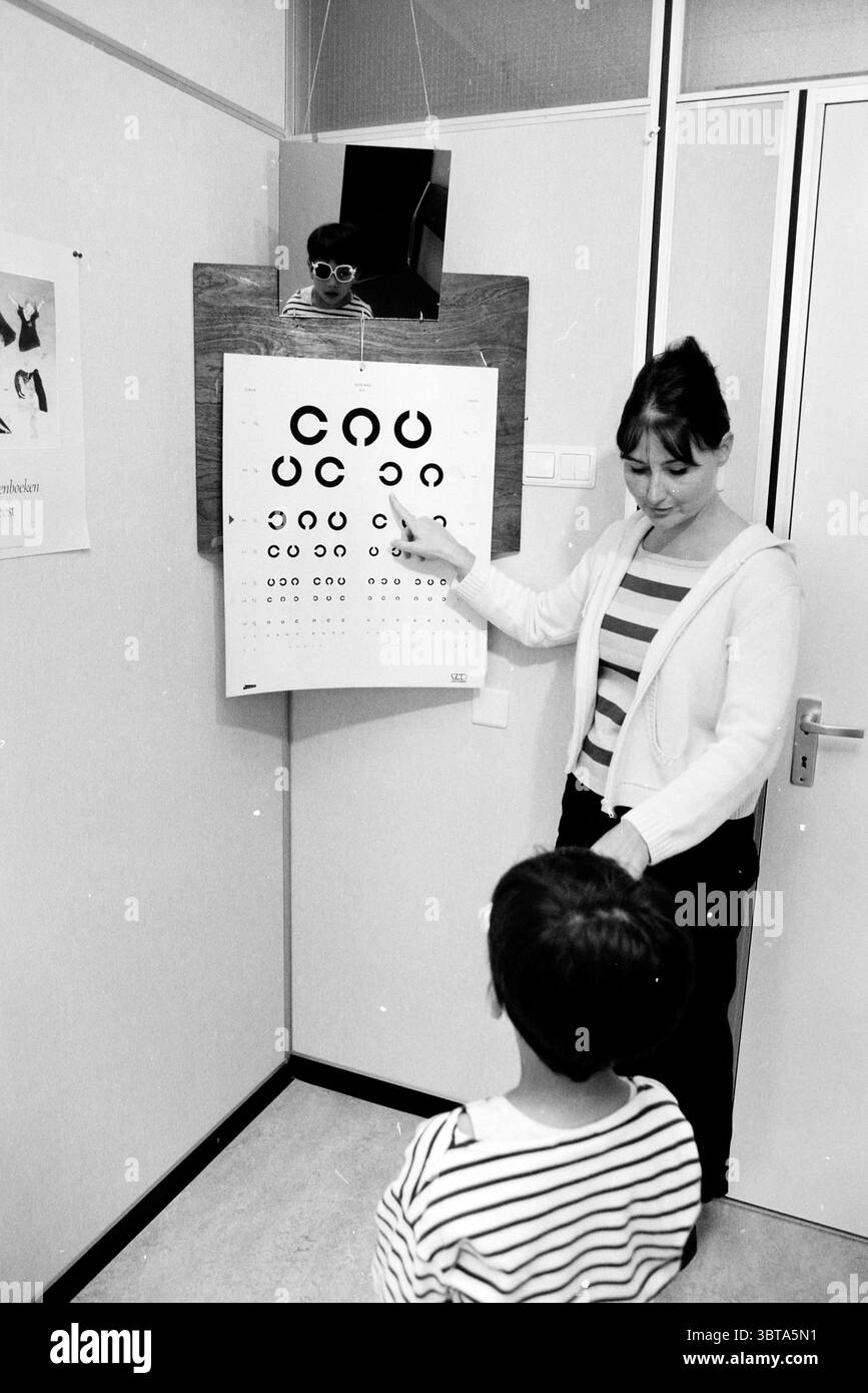 Examen de l'école de médecine de test oculaire, Whizgle News, Dutch Desk, pays-Bas, 1950 - 2000 sur 06-1999. Voici les rubriques de l'image. La scène met en scène une femme interagissant avec un enfant dans un environnement clinique ou éducatif. Elle se tient debout sur un côté, faisant des gestes vers un grand graphique qui affiche diverses formes circulaires en noir et blanc. L'expression de la femme semble concentrée et engagée, expliquant probablement quelque chose d'important sur le graphique. L'enfant, assis devant elle, est habillé d'une chemise rayée et lève les yeux, apparemment attentif. L'atmosphère générale se sent sérieuse mais suppo Banque D'Images