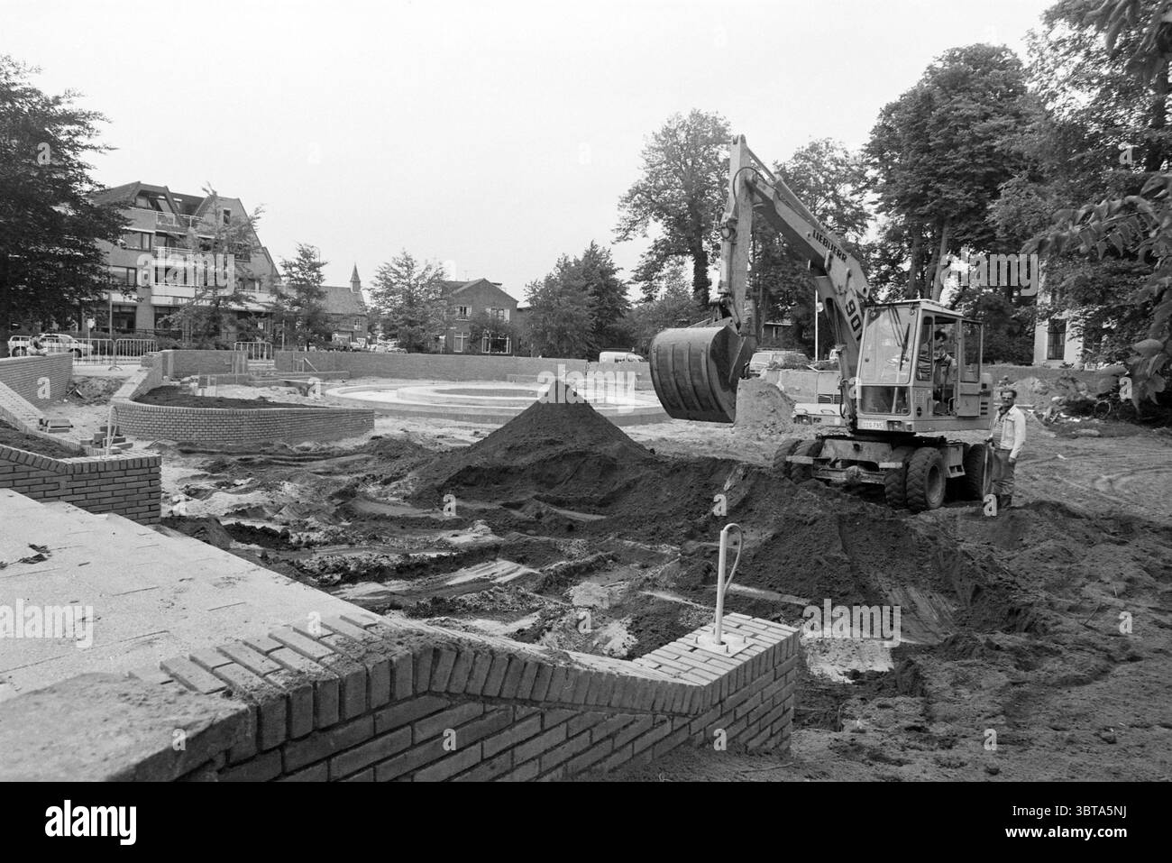 Construction étang jardin mairie Hillegom Hillegom Vijvers Hillegom, Whizgle News, Dutch Desk, pays-Bas, 1950 - 2000 on 18-07-1985. L'image montre ces rubriques. La scène représente un chantier de construction actif caractérisé par une quantité importante de terre étant déplacée autour. Une grande pelle, peinte dans des couleurs discrètes, domine le premier plan, son seau surélevé et posé au-dessus d'un monticule de sol brun rougeâtre. Les détails de la machine, tels que les tuyaux hydrauliques et les bras mécaniques, évoquent un sentiment de force industrielle. En arrière-plan, une structure circulaire partiellement construite est beginni Banque D'Images