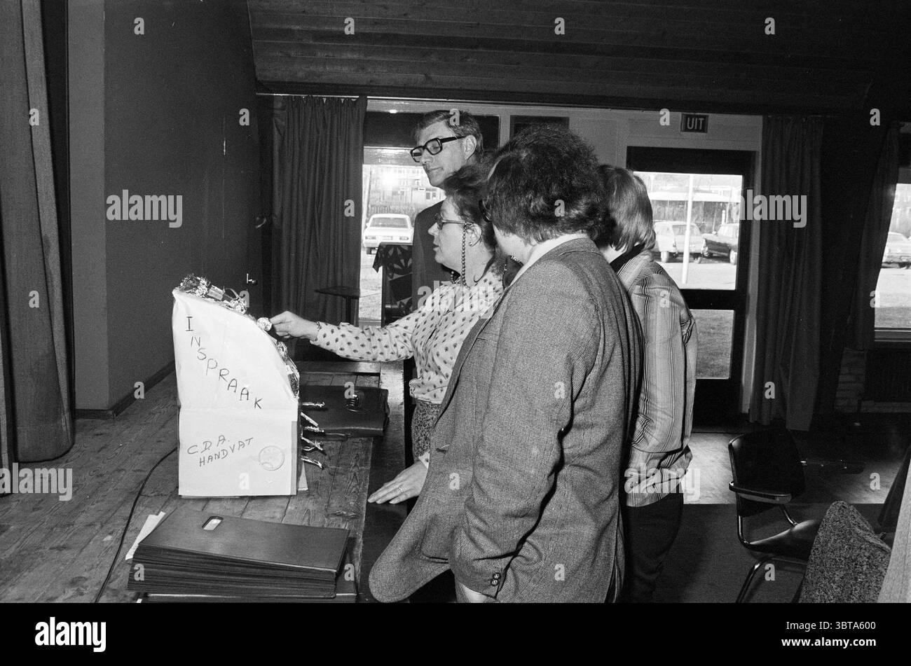 Budget municipalité de Heemskerk dans le Vrijburgt Conseil municipal de Heemskerk réunions, Whizgle News, Dutch Desk, pays-Bas, 1950 - 2000 le 08-12-1975. L'image contient ces rubriques. Dans une pièce faiblement éclairée, plusieurs individus sont rassemblés autour d’une table encombrée de dossiers et de papiers. Le cadre semble être une réunion communautaire ou un forum de discussion, avec un plafond en bois suggérant une atmosphère confortable et sérieuse. Quatre personnes se tiennent étroitement ensemble, se concentrant intentionnellement sur un objet au centre de la table. Leurs expressions véhiculent un mélange de curiosité et de concentration. Banque D'Images