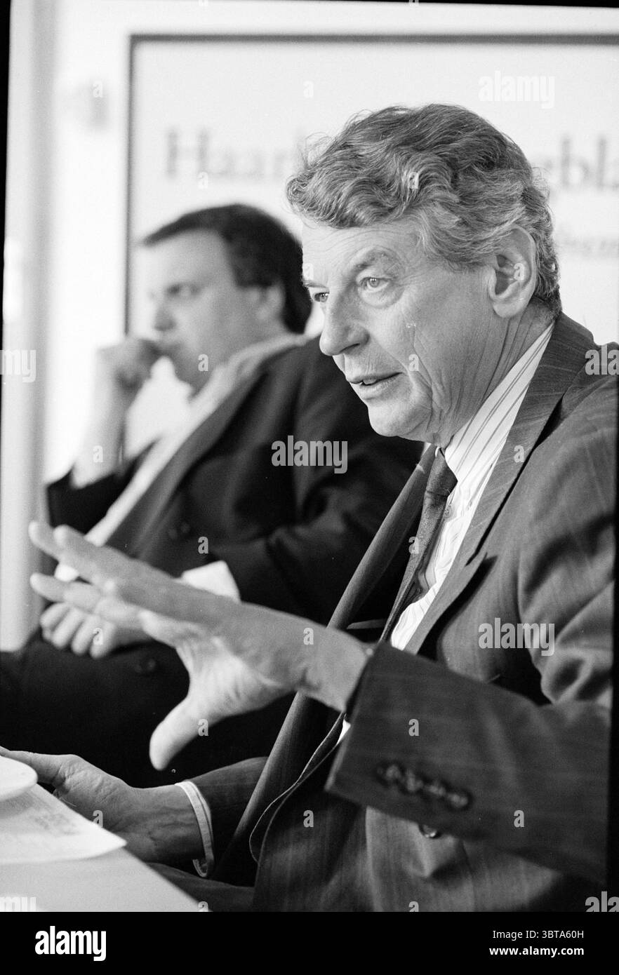 Interview] Kok + Rottenberg, Whizgle News, Dutch Desk, pays-Bas, 1950 - 2000 on 25-04-1994. L'image contient ces rubriques. Dans la scène, deux individus sont engagés dans une discussion. Le premier plan représente un homme aux cheveux gris argenté, portant un costume à fines rayures foncées, une chemise rayée et une cravate. Son expression transmet un sentiment d’autorité et d’engagement lorsqu’il fait des gestes de sa main droite, illustrant un point avec la paume ouverte et les doigts légèrement écartés. La lumière tombe sur son visage, accentuant les détails de ses traits et projetant des ombres douces qui mettent en valeur son expression. À l'arrière Banque D'Images